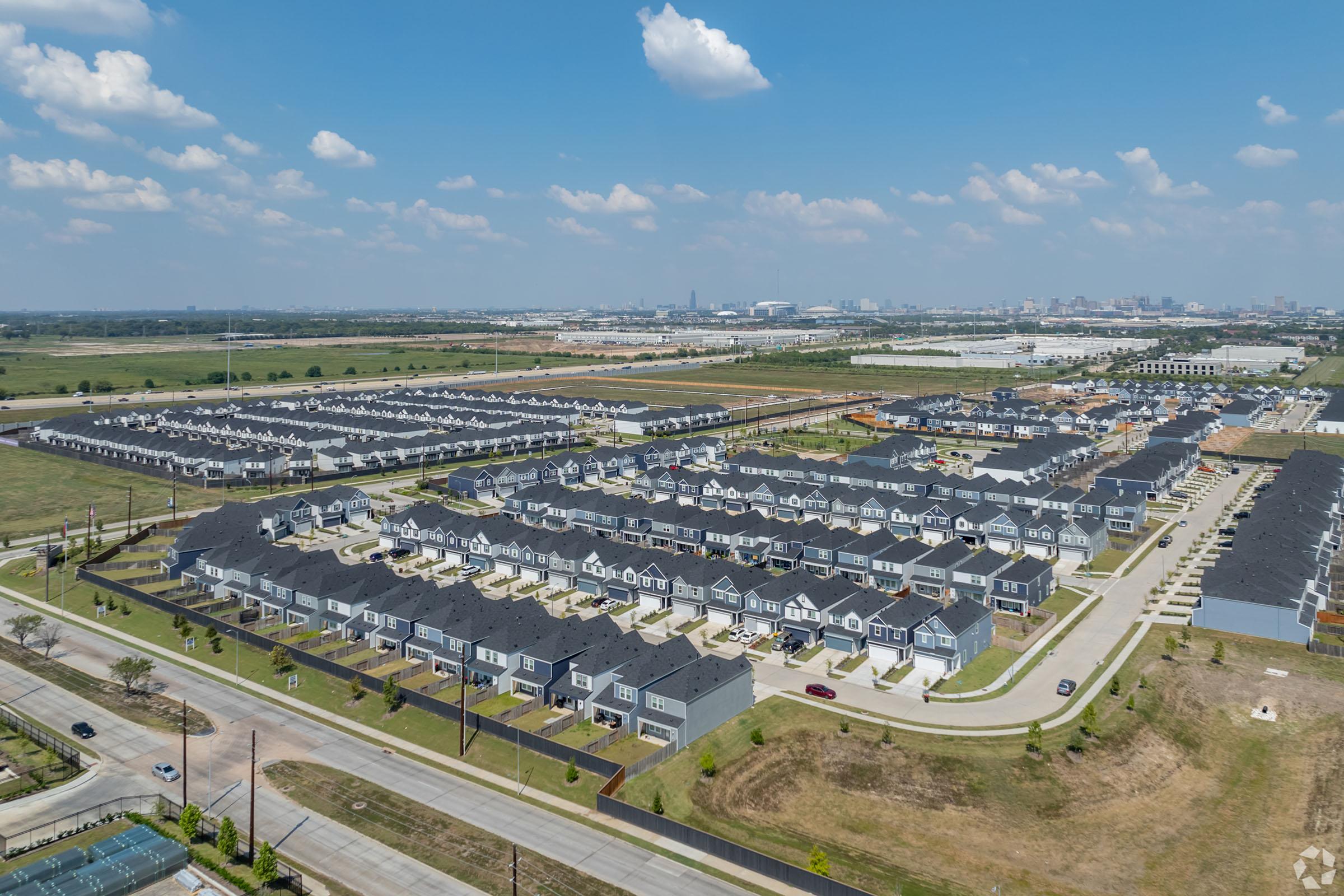 Aerial view of a residential neighborhood featuring rows of houses with dark roofs, surrounded by green fields and open spaces. In the distance, a city skyline is visible under a clear blue sky with scattered clouds. Roads and pathways connect the homes, showcasing a suburban layout.