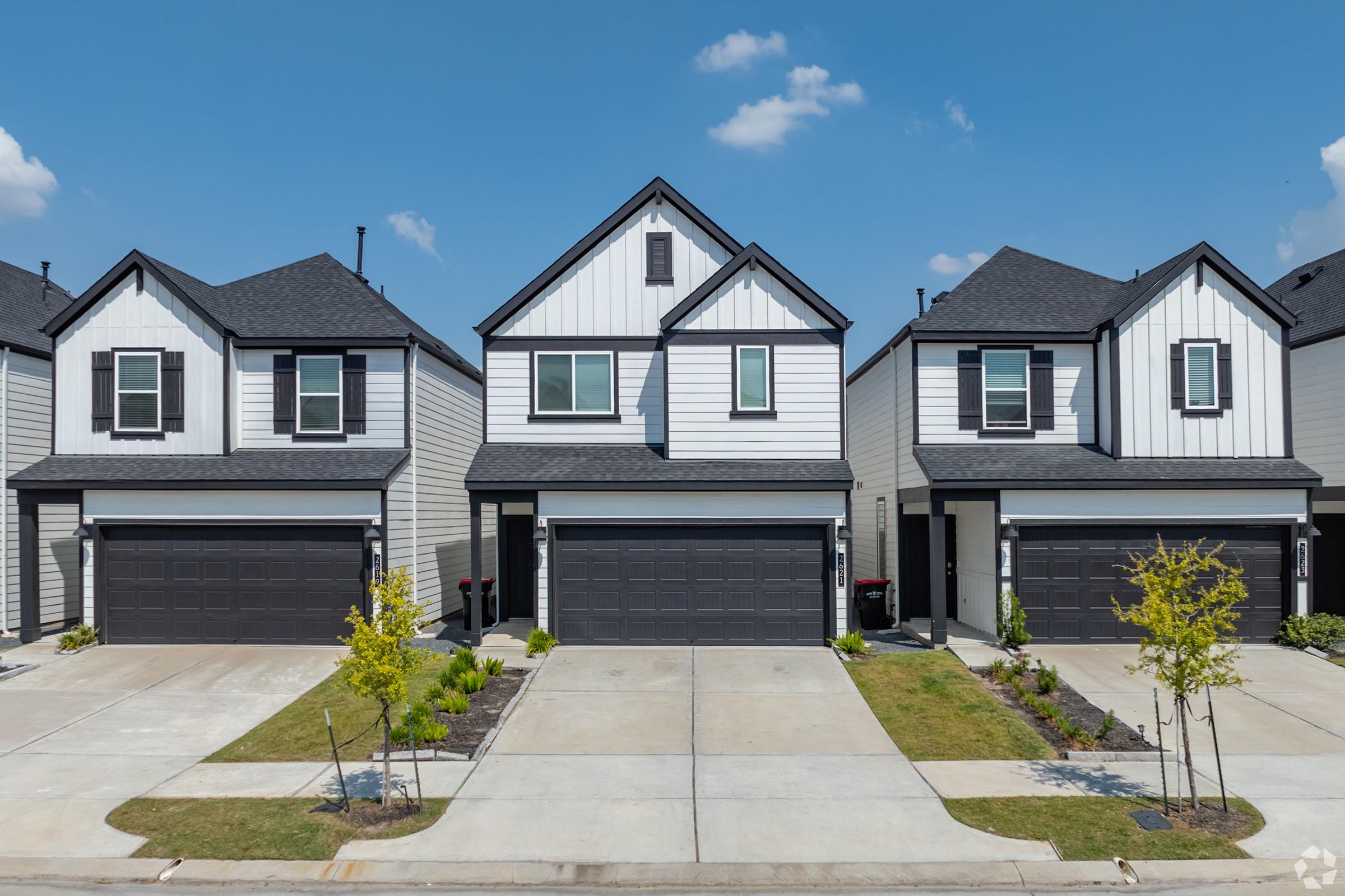 A row of modern, two-story homes featuring gray siding and black roofs. Each house has a garage and a small front yard with neatly trimmed grass and landscaping. The sky is clear with a few clouds, creating a bright and sunny atmosphere.