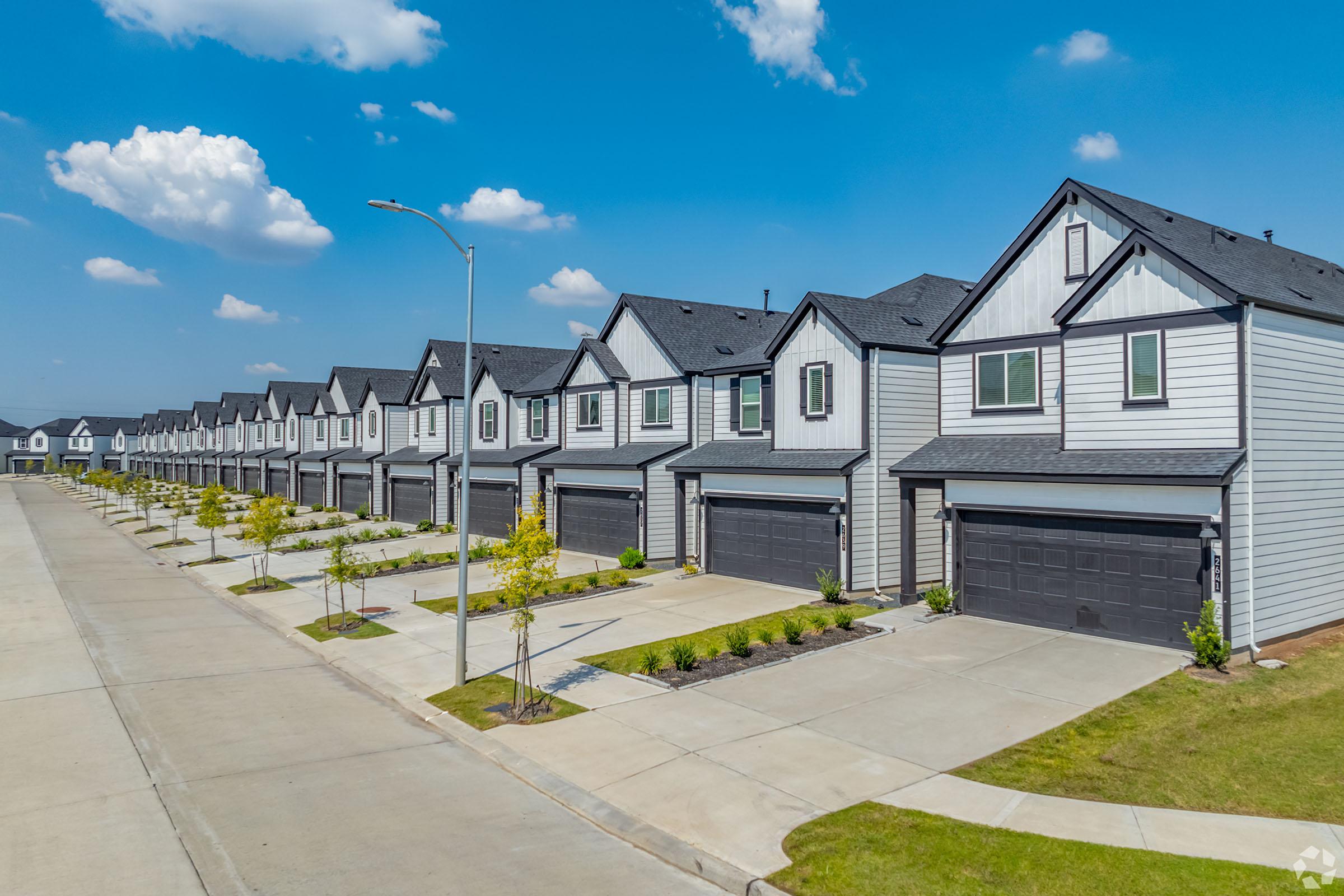 Row of modern townhouses with grey siding and black roofs, lined along a paved street. Each unit features a garage and small landscaped area with young trees. The sky is bright blue with scattered clouds, creating a sunny atmosphere.