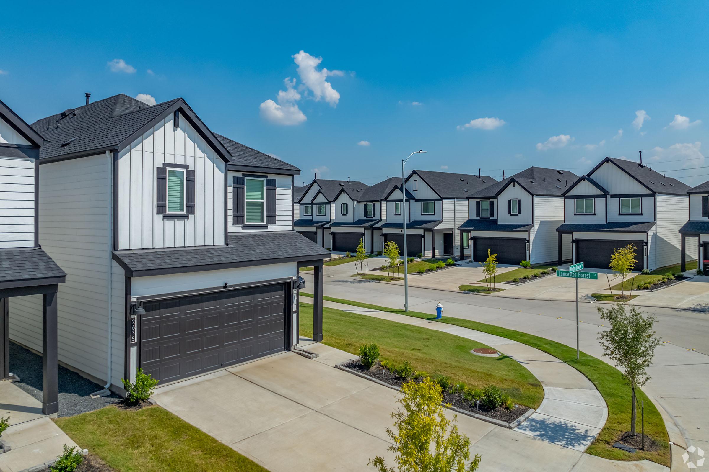 A modern residential neighborhood featuring two-story houses with black roofs and white exteriors. The homes are arranged in a grid layout, with well-maintained yards and small trees. A curved street is visible in the foreground, creating a welcoming suburban atmosphere under a bright blue sky.