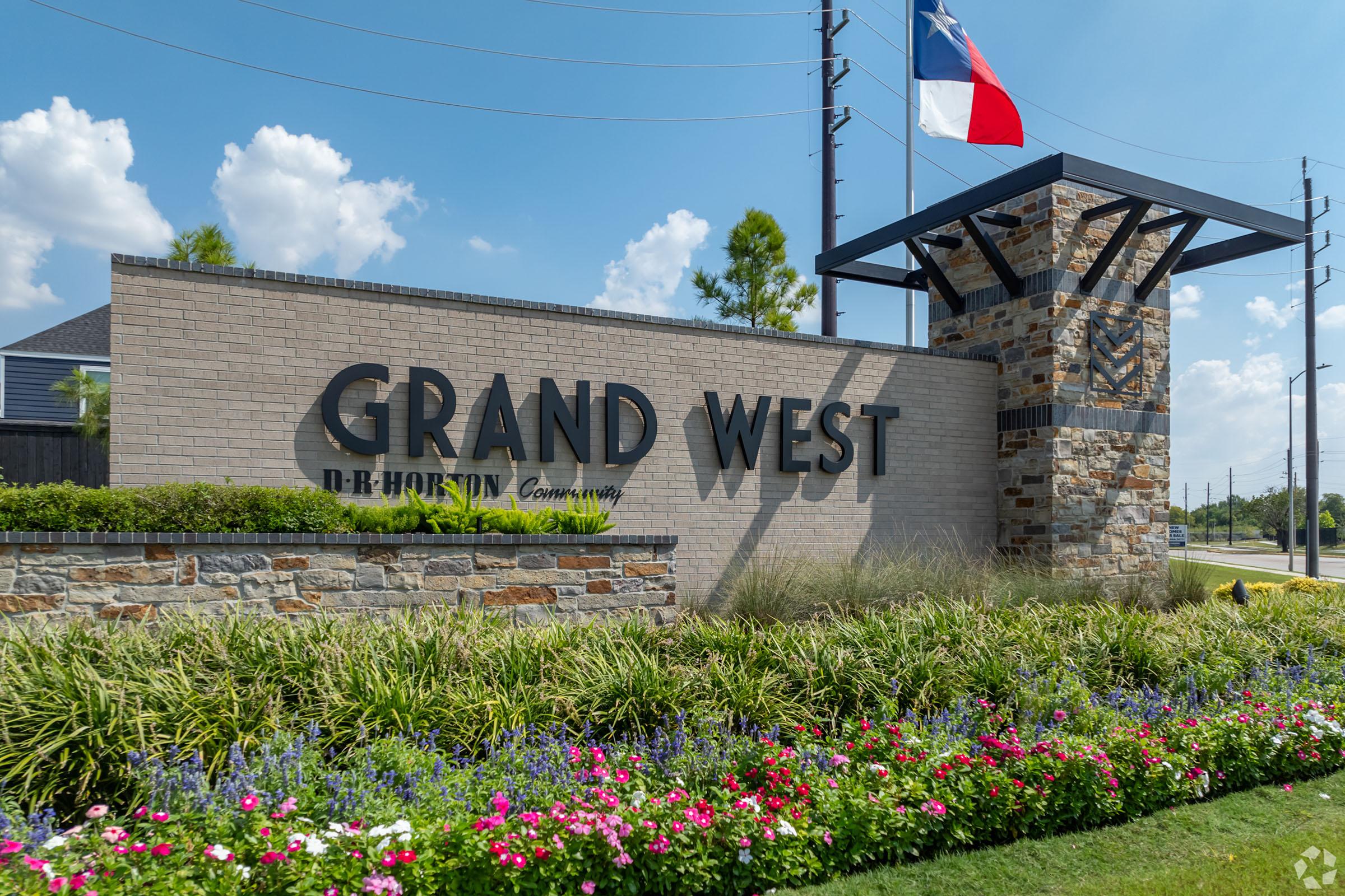 A stone and brick entrance sign for Grand West community, featuring bold lettering against a backdrop of colorful flowers and greenery. A Texas flag is prominently displayed at the structure, under a blue sky with scattered clouds.