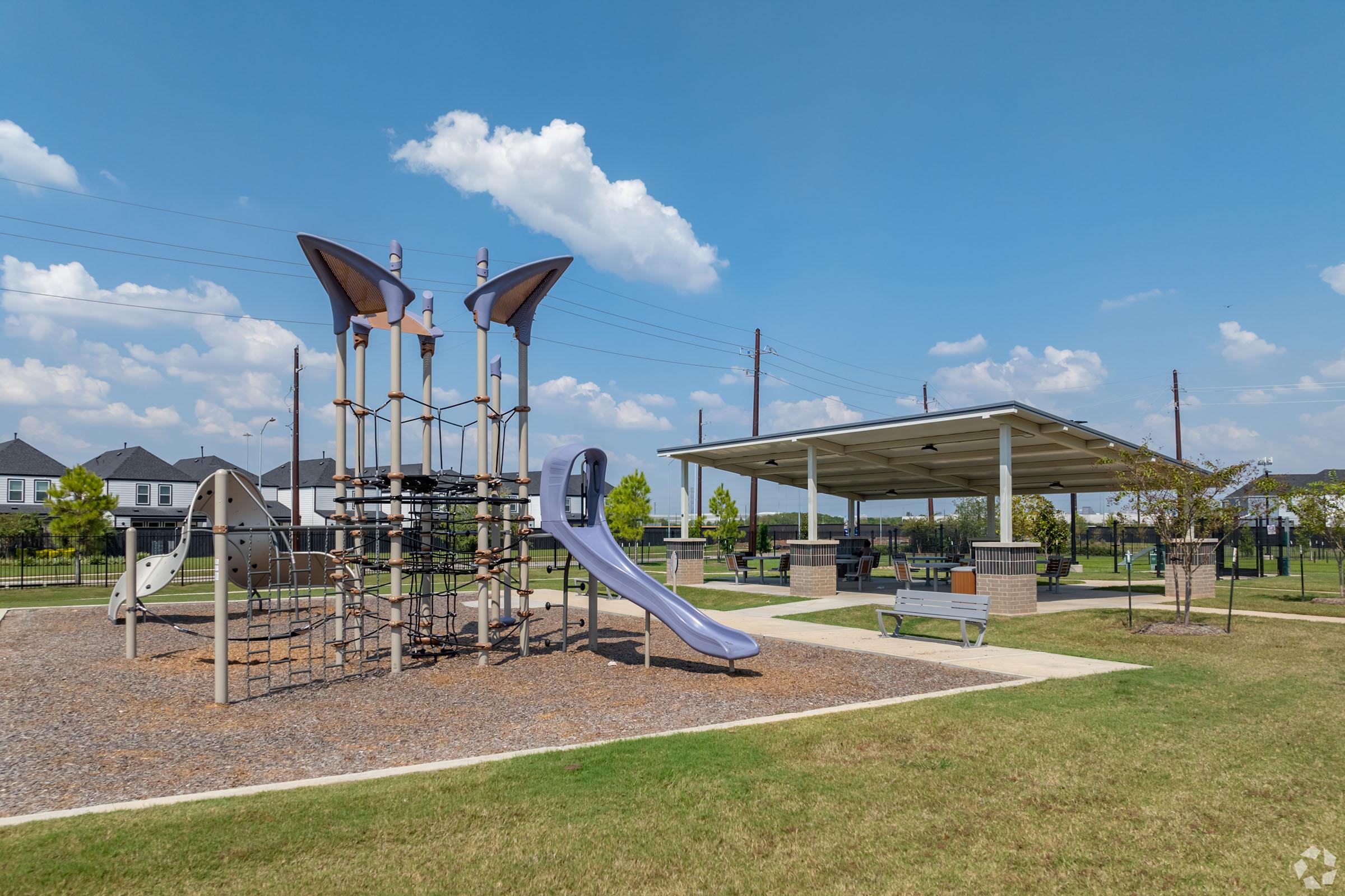 A sunny playground scene featuring a modern play structure with climbing frames and slides. Nearby, there's a shaded picnic area with benches, surrounded by green grass and a clear blue sky with fluffy white clouds. Residential buildings are visible in the background.