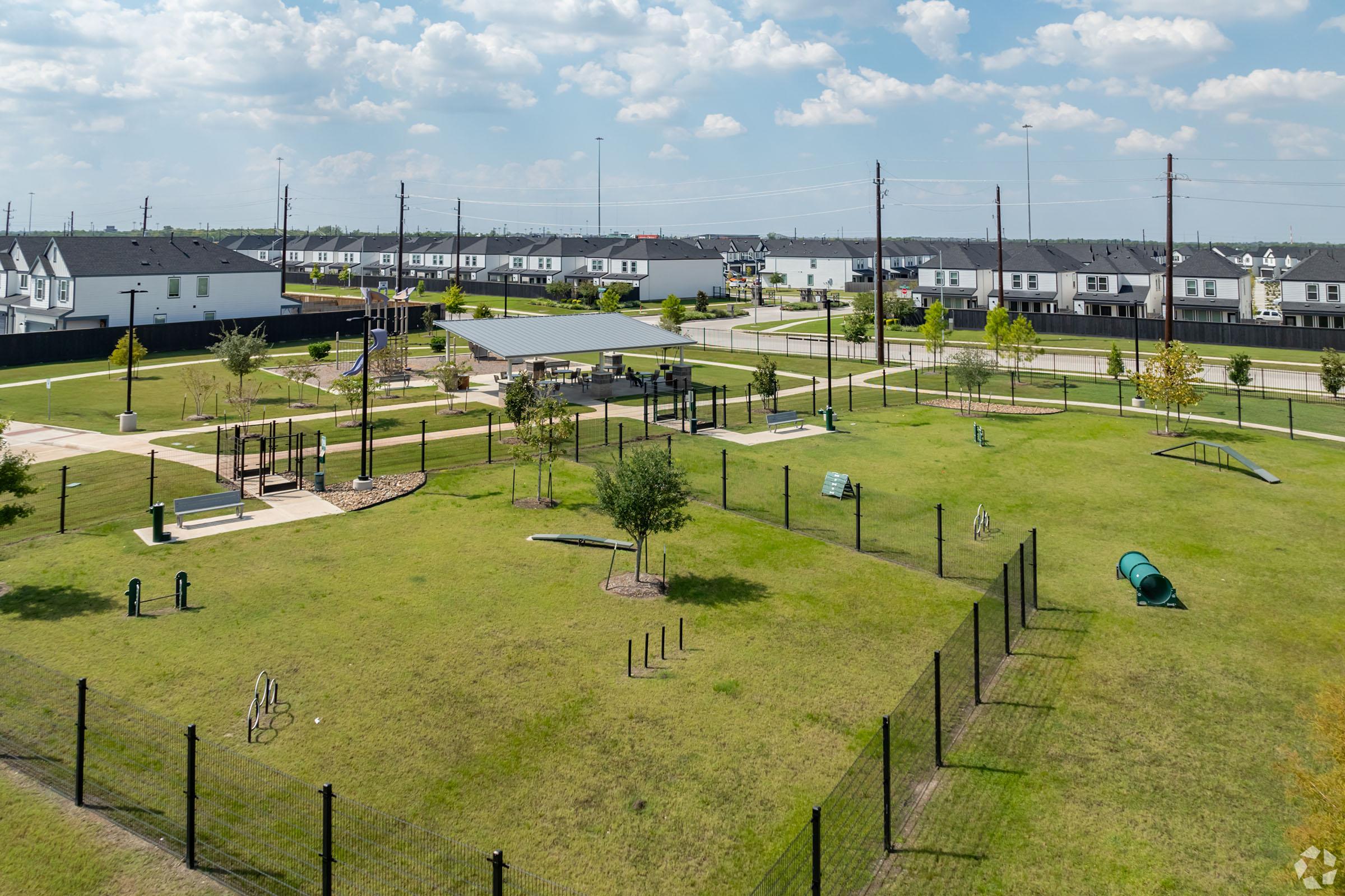 Aerial view of a well-maintained park featuring green grass, a variety of playground equipment, and exercise stations. The park is surrounded by a black fence, with residential buildings in the background under a blue sky with scattered clouds. Smooth pathways are visible among the landscaped areas.
