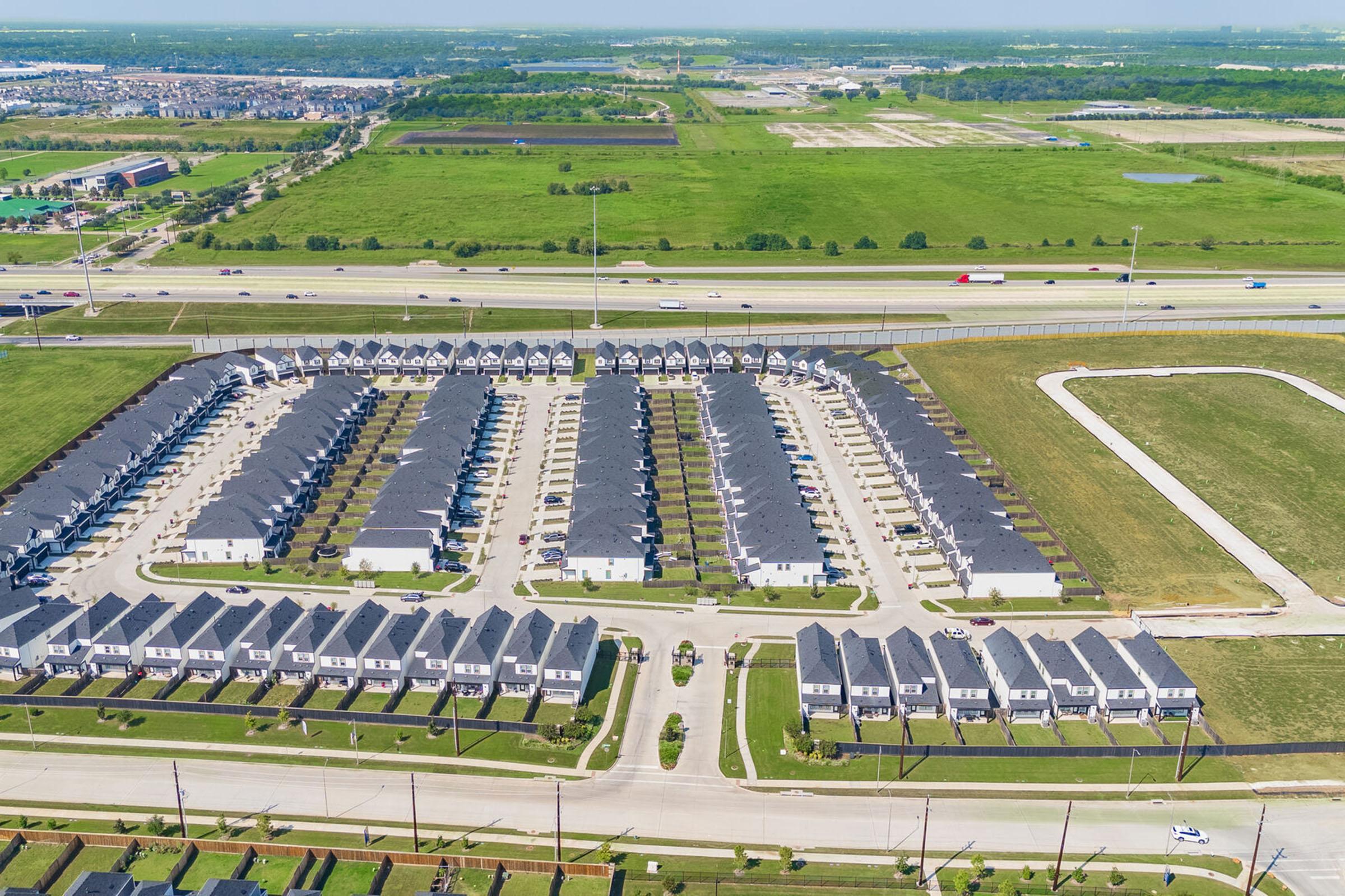 Aerial view of a residential neighborhood featuring multiple rows of houses arranged in a grid pattern, with well-maintained lawns and driveways. Surrounding fields and open land are visible in the background, along with a highway and some light traffic. Clear skies and greenery create a spacious, suburban atmosphere.