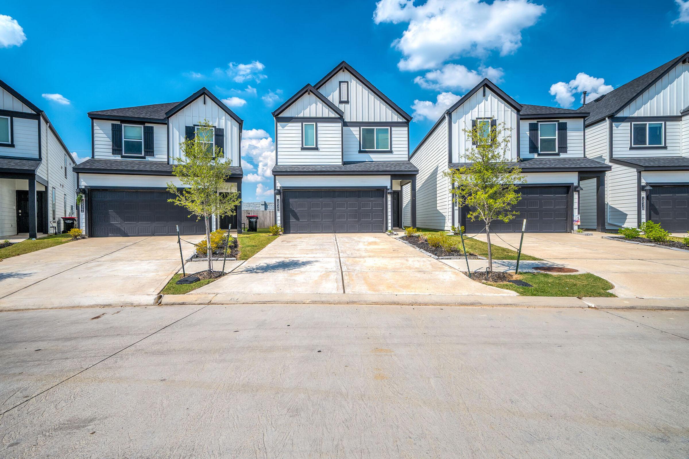 A row of contemporary two-story houses with gray siding and garages, set against a bright blue sky with fluffy clouds. Small trees and landscaped patches line the driveway. The scene conveys a suburban residential area, showcasing well-maintained homes with ample outdoor space.