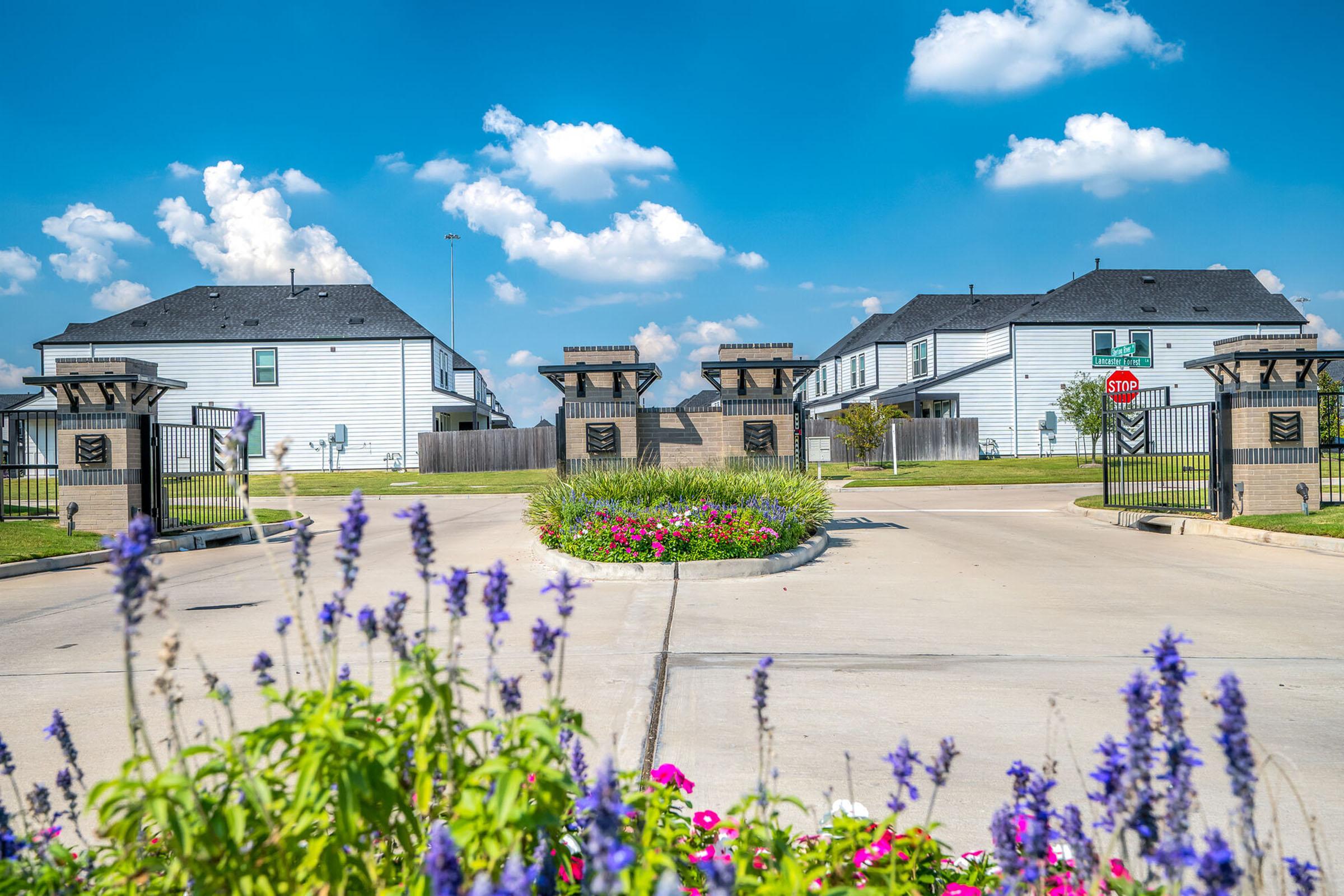 A well-maintained entrance to a residential community featuring a landscaped roundabout with colorful flowers in the foreground. Two decorative gates mark the entrance, flanked by modern homes under a bright blue sky with scattered clouds. A stop sign is visible near the gates.