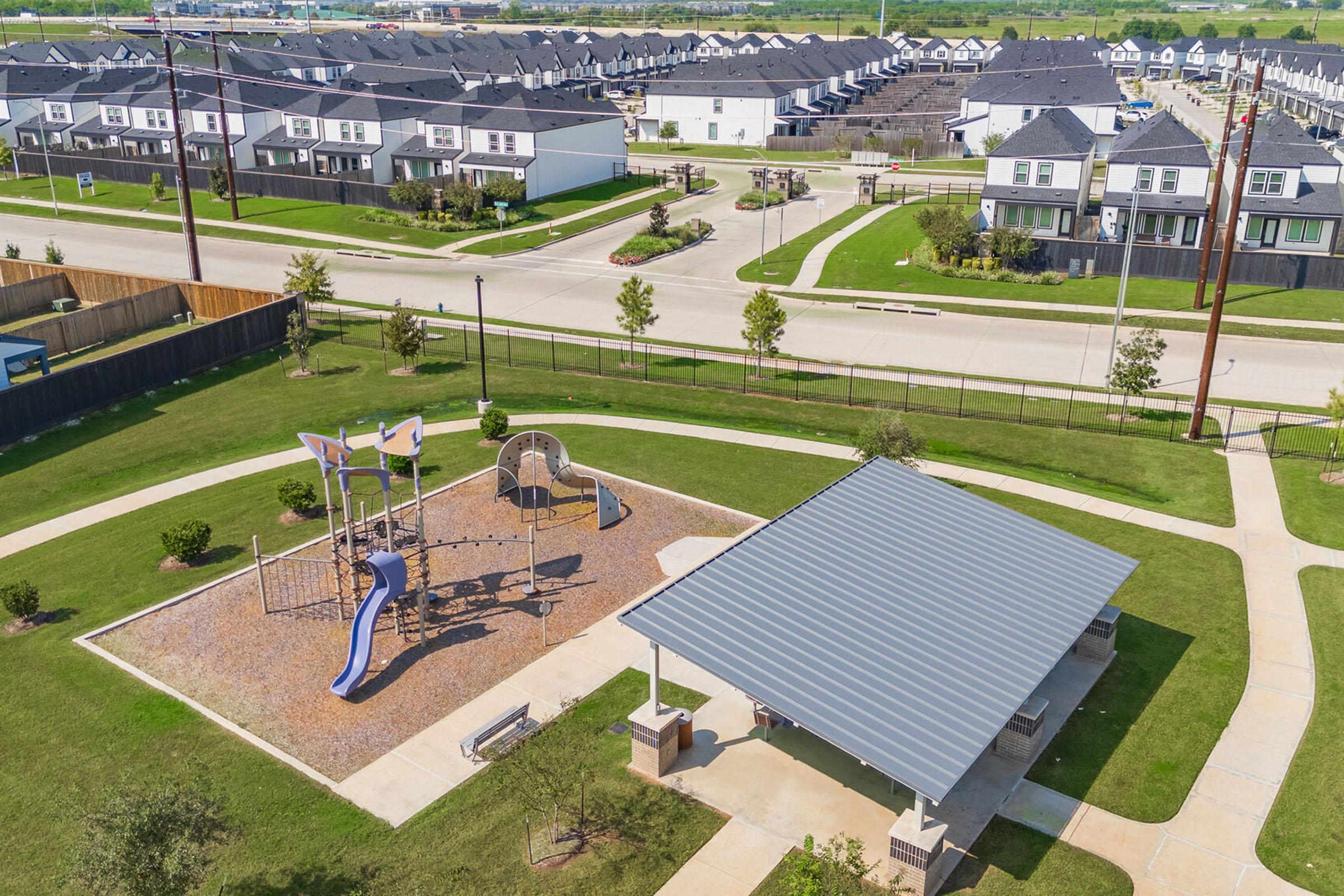 Aerial view of a community park featuring a playground with slides and climbing structures, surrounded by green grass. There is a shaded pavilion nearby, and in the background, rows of residential houses can be seen along a quiet street. The scene is bright and sunny, depicting a welcoming neighborhood.