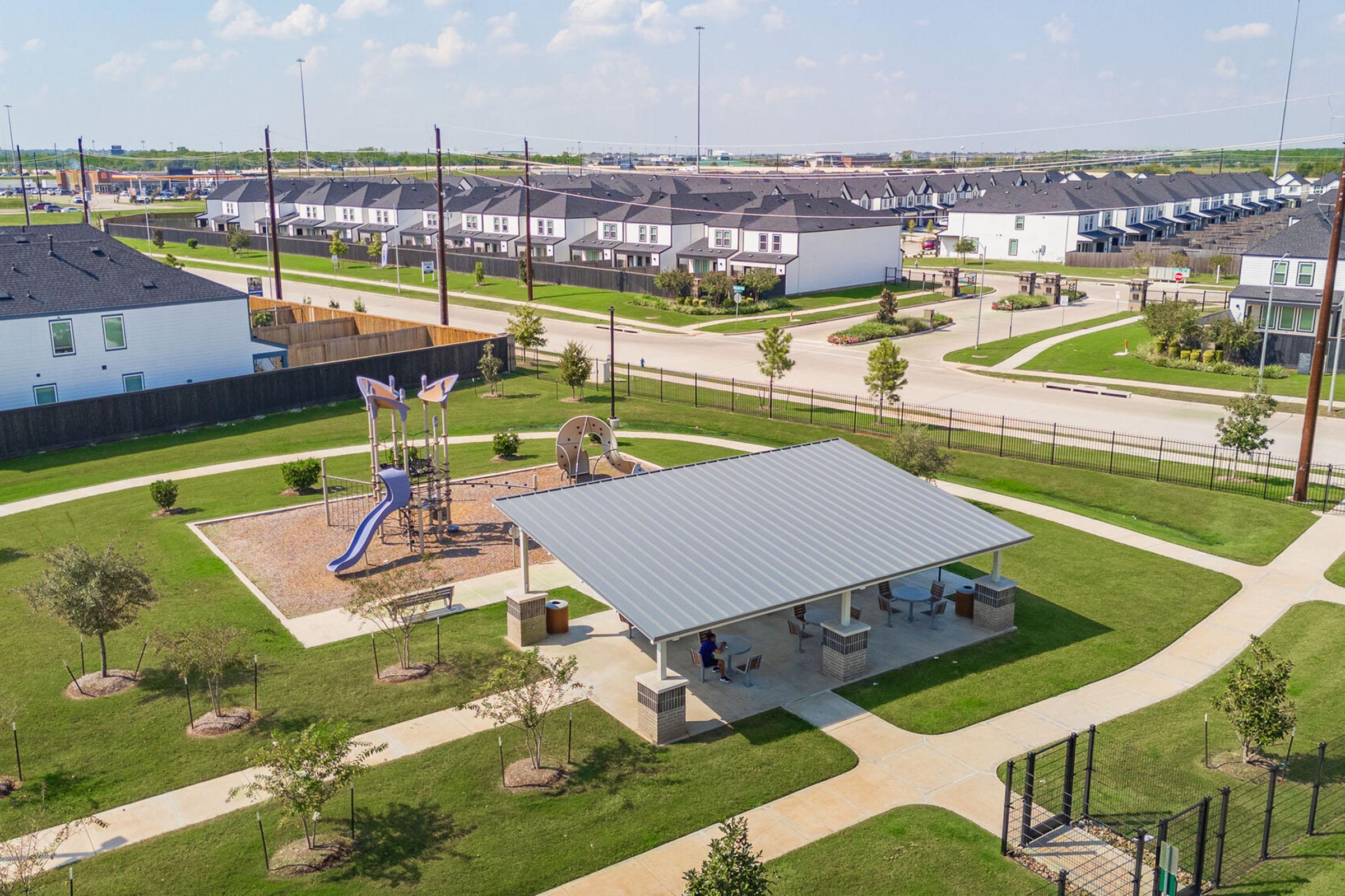 Aerial view of a community park featuring a playground with a blue slide, a picnic area with a roof, and landscaped greenery. In the background, there are residential buildings and a street lined with trees, under a bright blue sky with a few clouds.