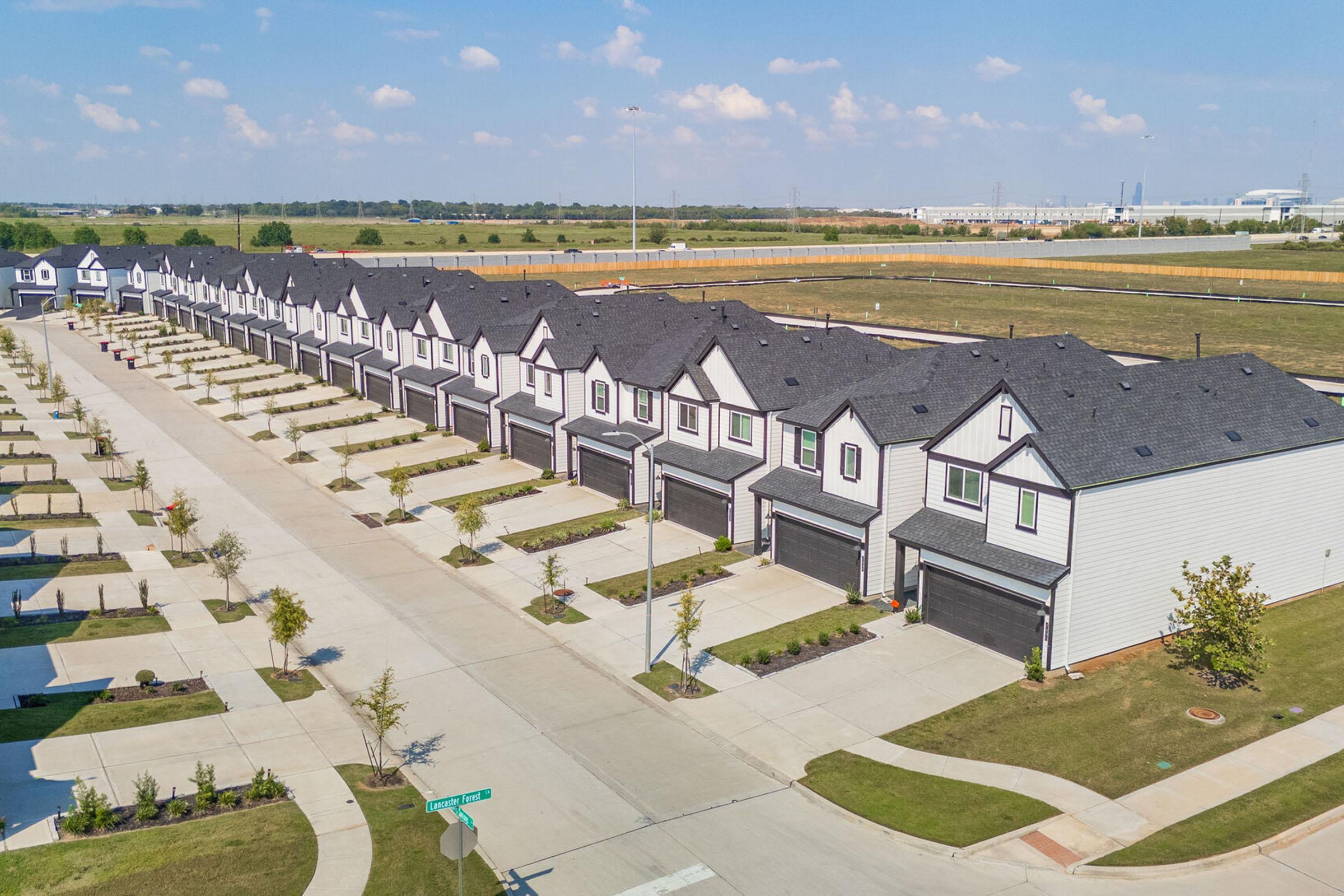 Aerial view of a suburban neighborhood featuring rows of identical two-story houses with dark roofs and garages. The street is lined with neatly maintained sidewalks and landscaping, while open fields are visible in the background under a clear blue sky.