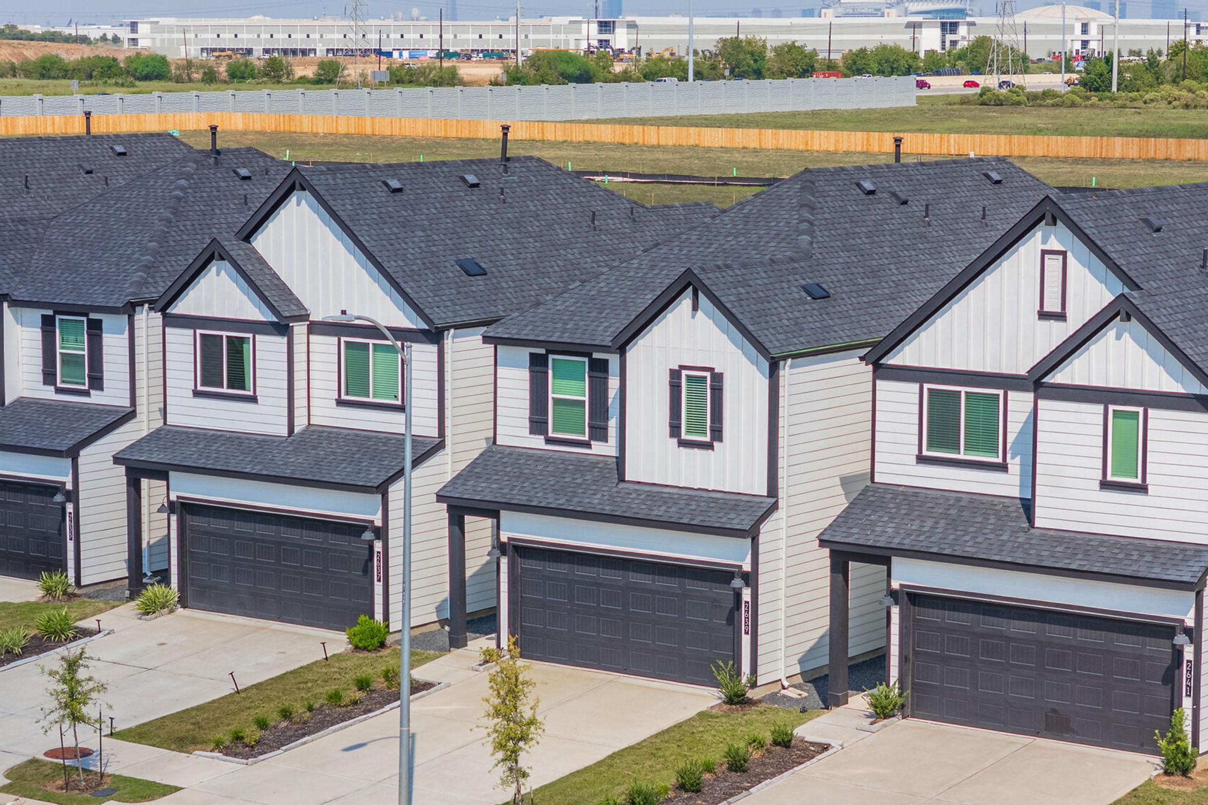 A row of newly constructed homes features modern architecture with gray siding and dark roofs. Each house has a driveway, green windows, and landscaped front yards. In the background, a vast open space and industrial buildings are visible, suggesting a suburban setting.