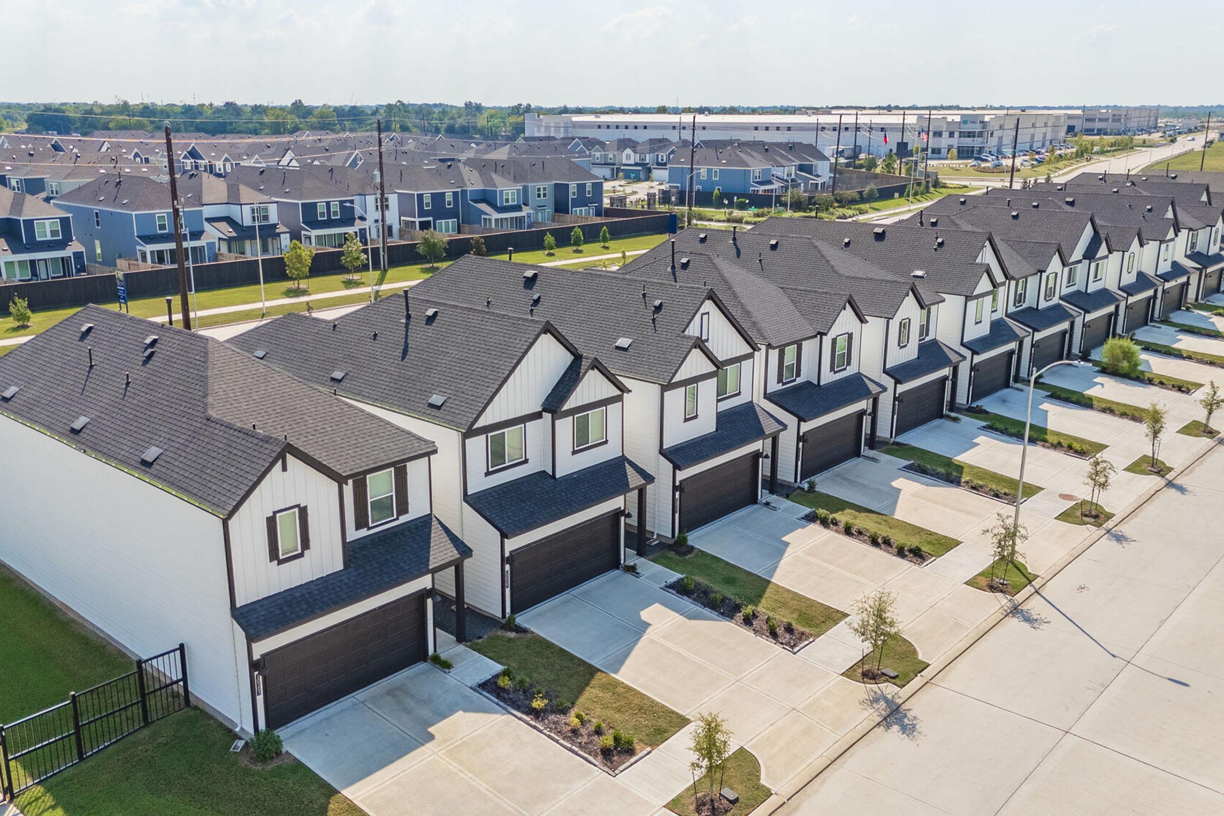 Aerial view of a modern suburban neighborhood featuring rows of stylish two-story homes with black roofs and white exteriors. Well-maintained lawns and driveways are visible, along with nearby streets lined with additional houses in the background. Clear blue sky adds to the bright atmosphere.