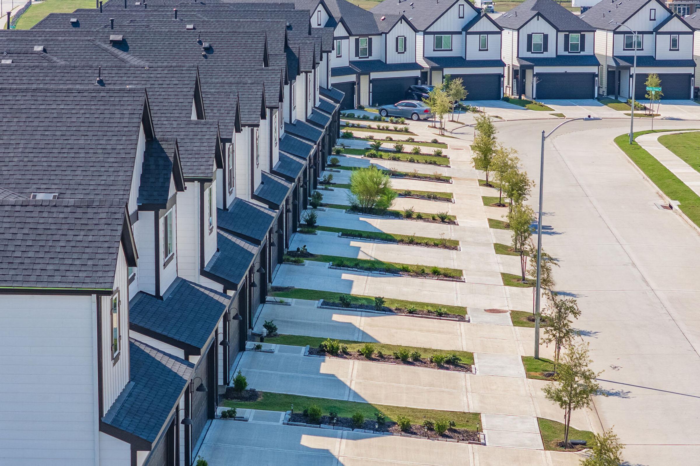 Aerial view of a row of modern townhouses with black roofs and white exteriors. The driveways are lined with neatly trimmed grass and small trees, and the street is quiet with a few parked cars in the distance. Bright sunlight casts shadows across the scene, creating a clear and inviting atmosphere.