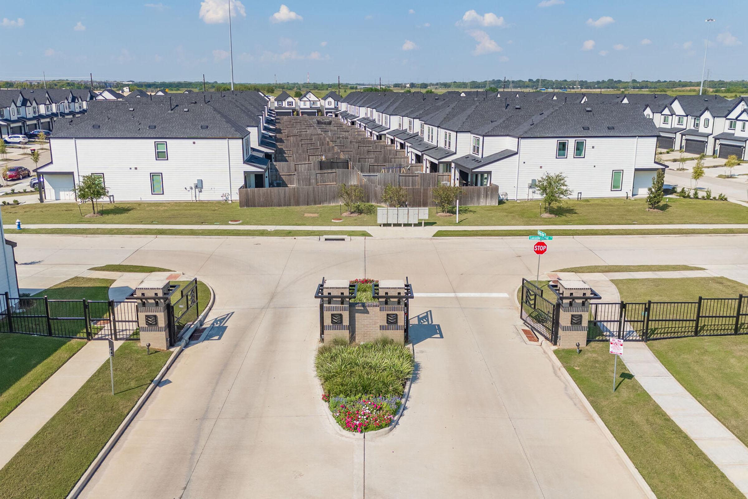 Aerial view of a residential community entrance featuring a gated entry with brick pillars. The road leads into a neighborhood with multiple houses visible, surrounded by green lawns and landscaping. Clear blue sky with a few clouds in the background.