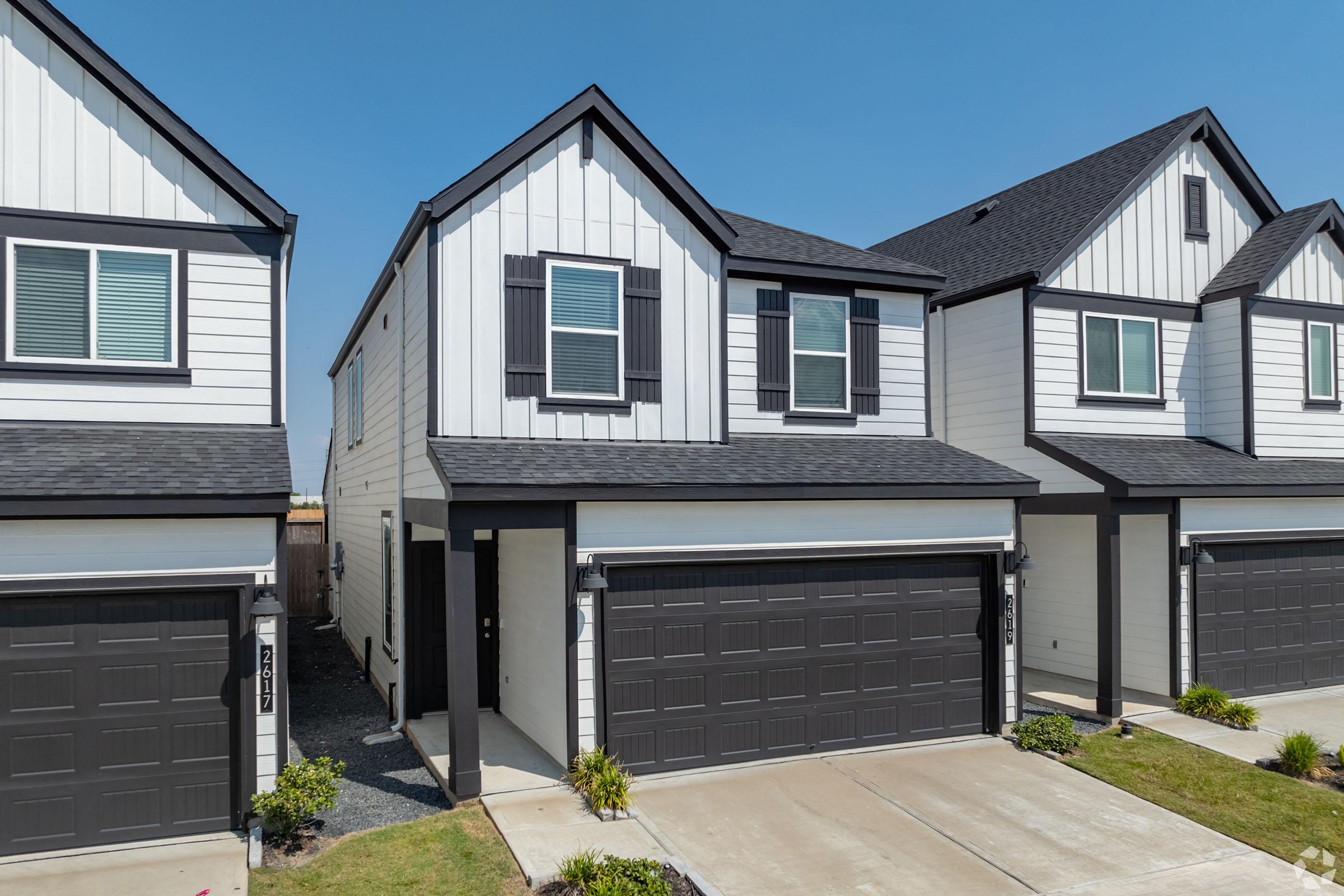 A modern two-story home with a symmetrical design, featuring white board and batten siding and black trim. The house has a dark roof, front porch, and a two-car garage. Neatly landscaped with small shrubs and a clear blue sky in the background. The adjacent homes are similar in style.