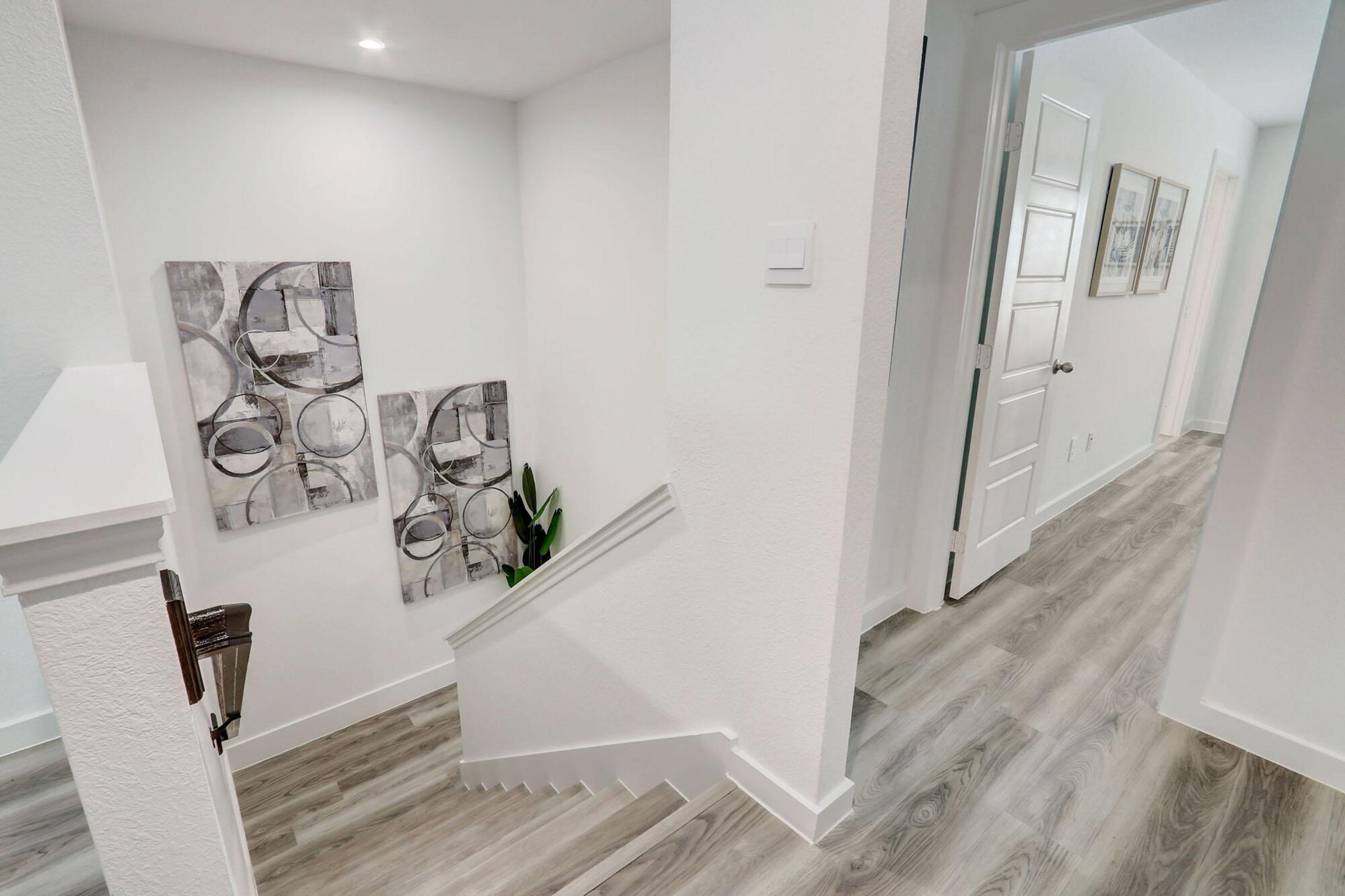 A well-lit, modern staircase with light-colored walls and wooden flooring. Decorative paintings featuring abstract designs are mounted on the wall beside the stairs. A door is visible at the end of the hallway, leading to another part of the home. Green plants add a touch of nature to the space.