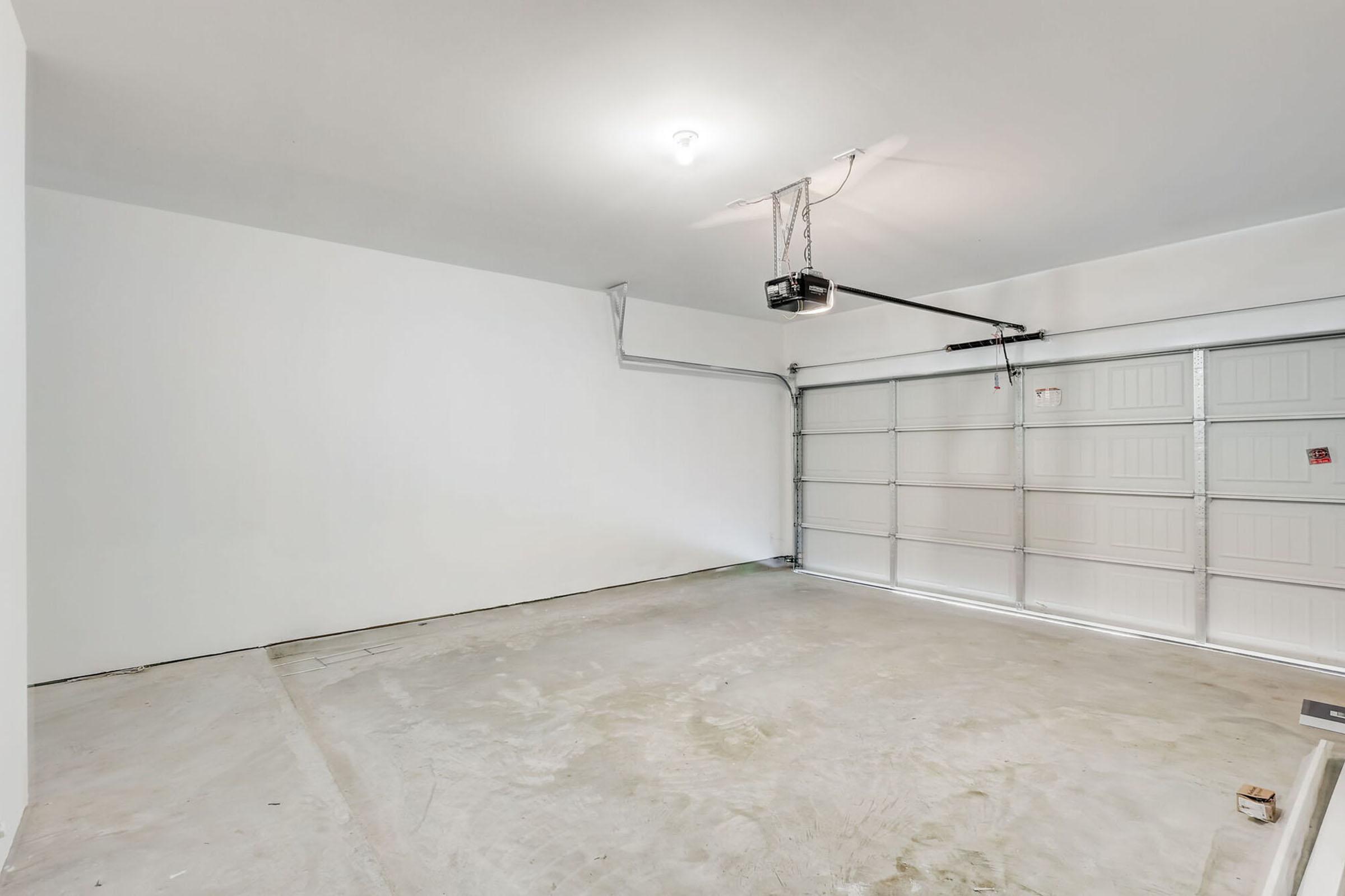 A spacious, empty garage with plain white walls and a concrete floor. The overhead garage door is partially open, revealing a simple mechanical door opener. The room has a light fixture on the ceiling, providing bright illumination. There are no vehicles or storage items present.