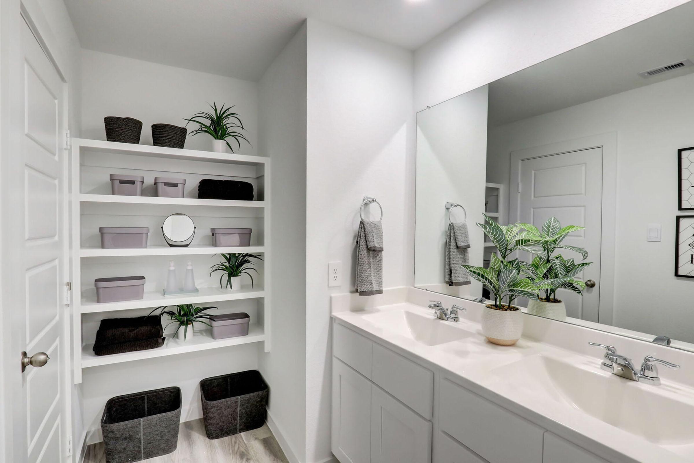 A modern bathroom featuring a double sink vanity with an elegant mirror above. The shelves are neatly organized with decorative baskets and plants. Two towels hang from hooks, and there are small storage bins on the floor, creating a clean and minimalist aesthetic. Bright and well-lit space with neutral colors.