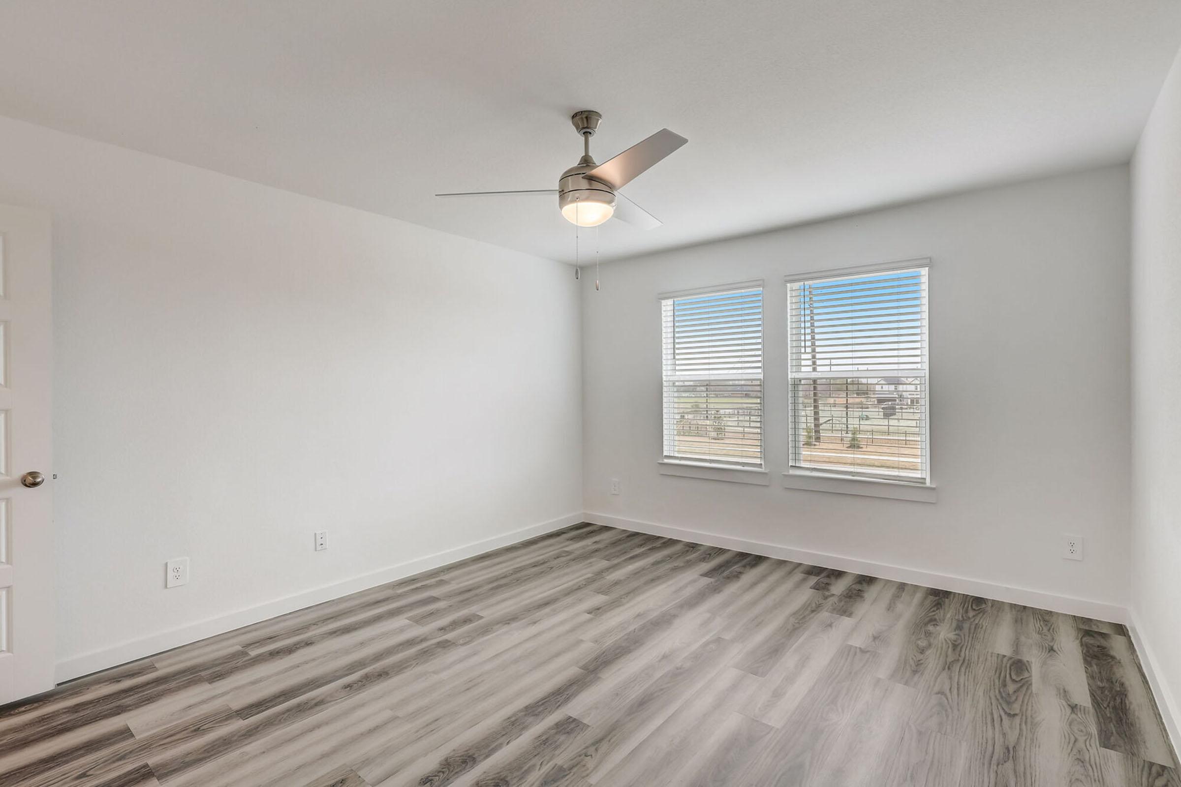 A modern, minimally decorated room with light-colored walls and wooden flooring. It features a ceiling fan and two windows with blinds, allowing natural light to enter. The space appears tidy and spacious, suitable for various uses, such as a bedroom or office.