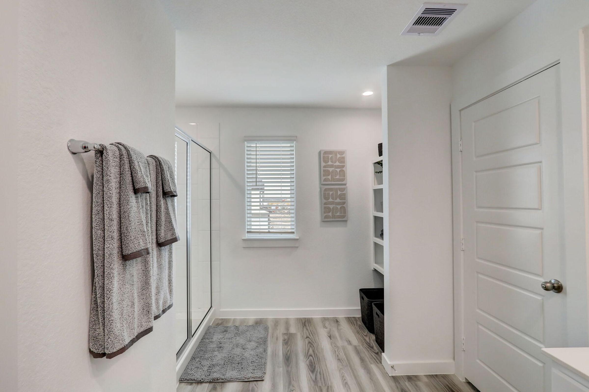 A modern bathroom featuring a glass shower, two gray towels hanging on a towel rack, a large white door, and a shelf with black storage bins. The space is bright and minimalistic, with light-colored walls and a wood-like floor, complemented by natural light from a window with blinds.