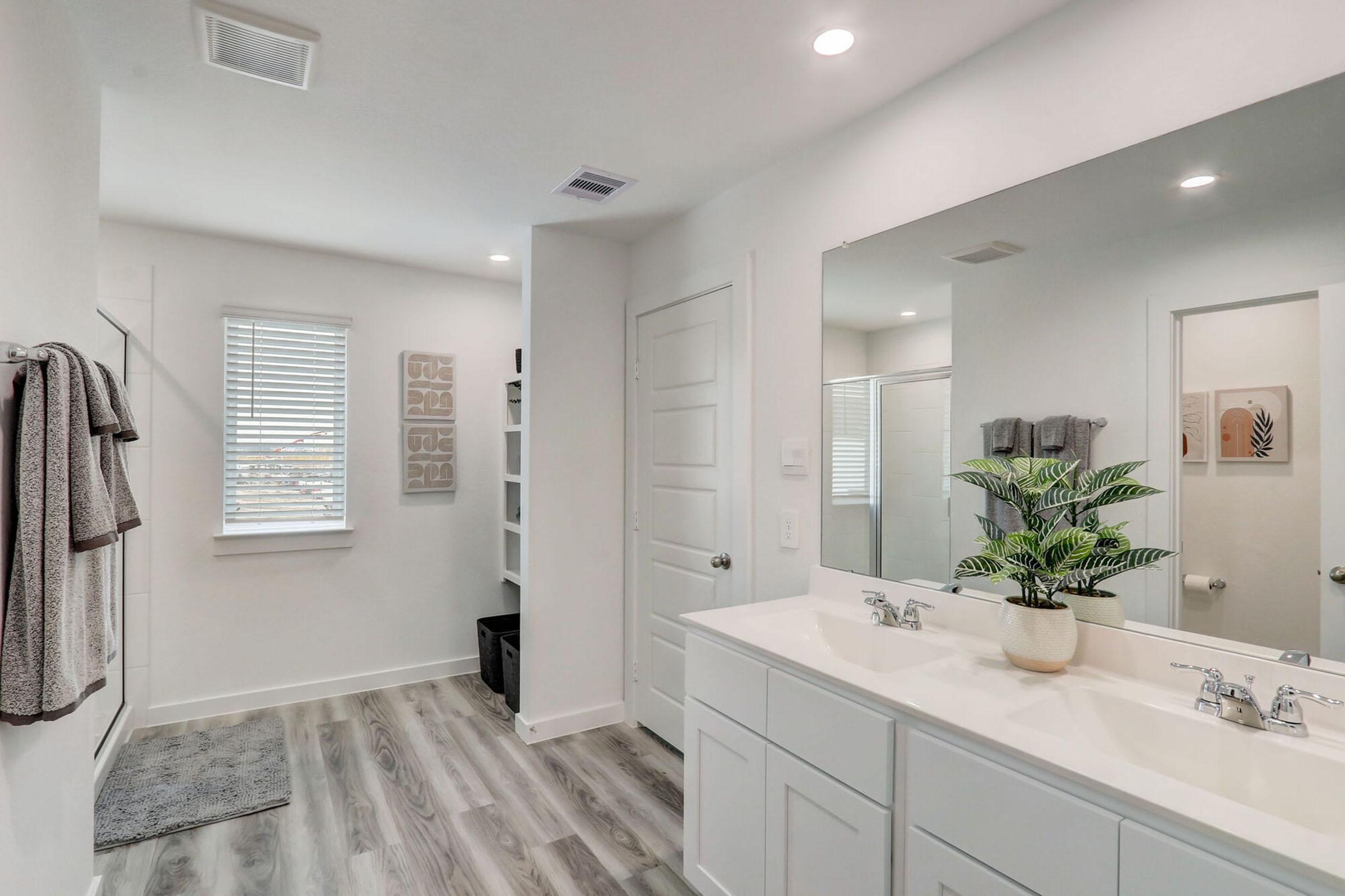 Bright and modern bathroom featuring double sinks, a large mirror, and a minimalist design. The room has grey-toned flooring, a window with blinds, and decorative wall art. A potted plant adds a touch of greenery, while neatly hung towels and organized shelves contribute to the clean aesthetic.