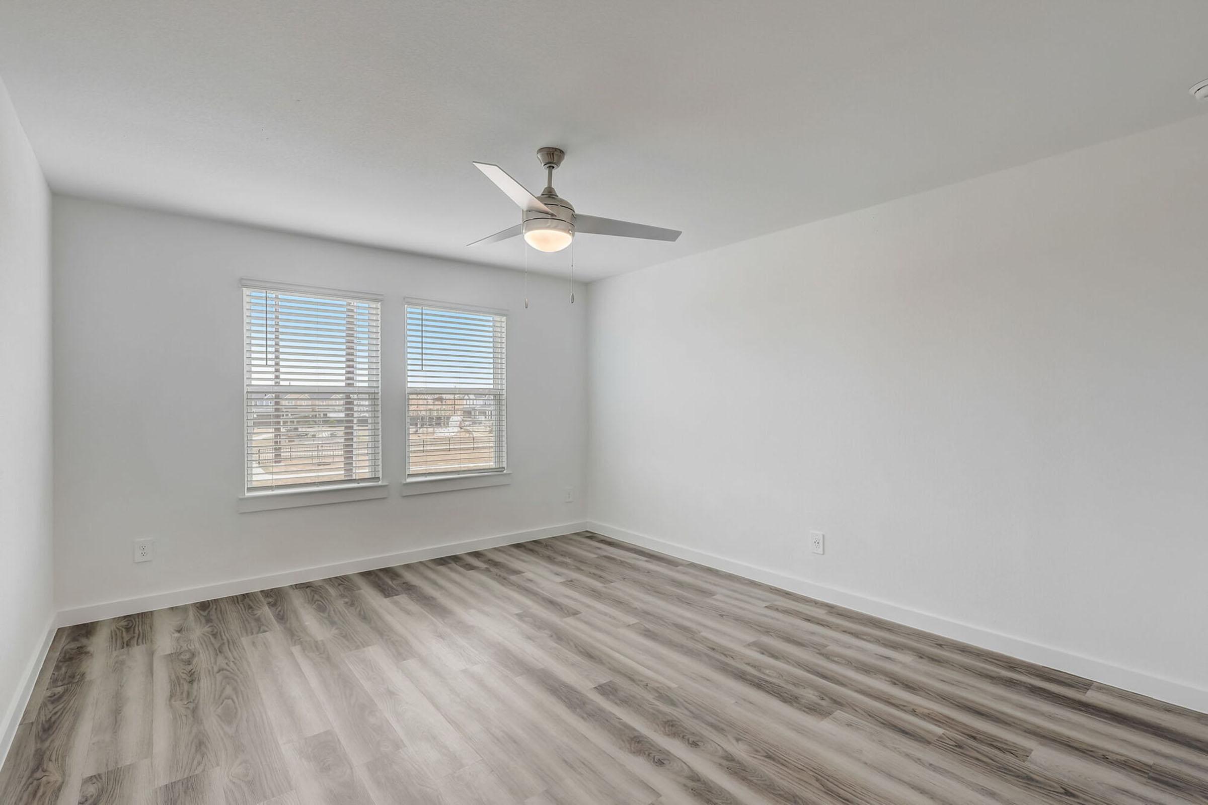 A bright, empty room featuring light gray walls and wooden flooring. It has a ceiling fan and two windows with white blinds, providing natural light. The overall atmosphere feels clean and modern, suitable for various interior design styles.