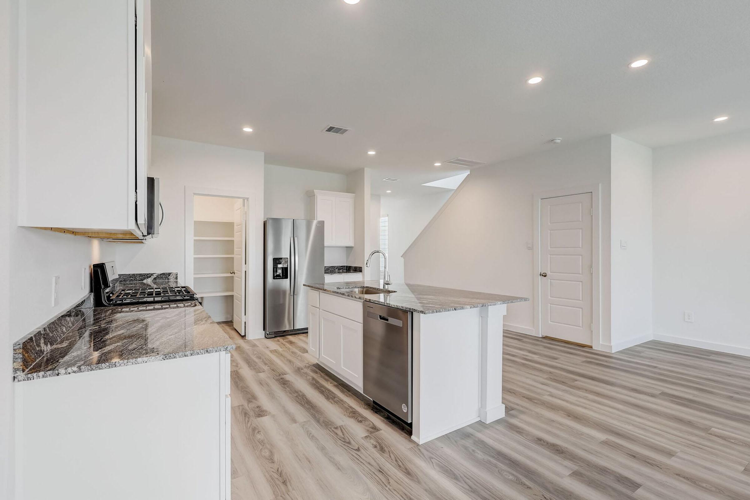 Modern kitchen featuring a central island with a granite countertop, stainless steel appliances including a refrigerator and dishwasher, white cabinetry, and an open layout with wood-like flooring. Natural light fills the space, highlighting the clean, contemporary design.