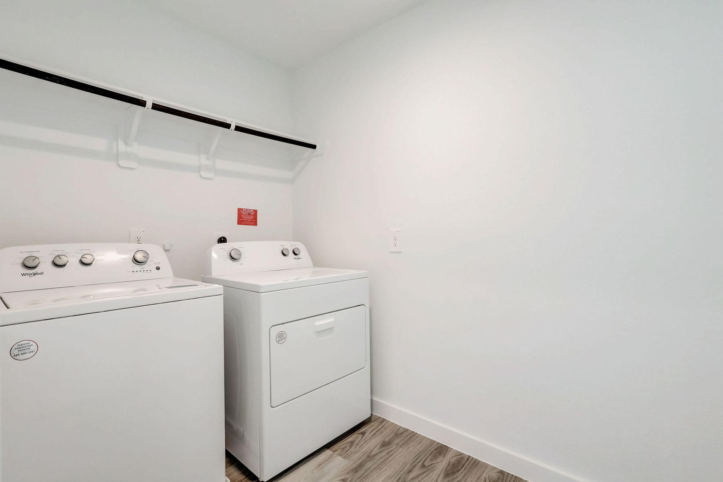 A laundry room featuring two white appliances: a washing machine and a dryer. The appliances are positioned against a light-colored wall, with a shelf above for storage. The flooring is light wood, enhancing the airy and clean atmosphere of the space.