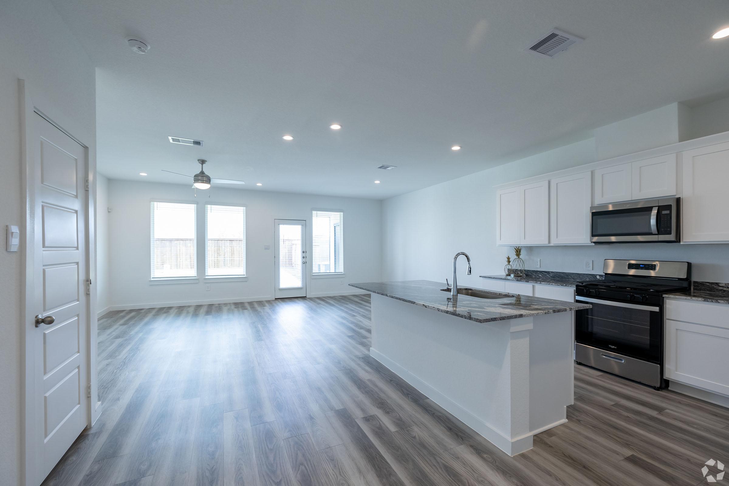 A modern, bright open-concept living space featuring a kitchen island with a granite countertop, stainless steel appliances, and white cabinetry. Large windows fill the room with natural light, showcasing a spacious layout with wood-style flooring and a door leading to an outdoor area.