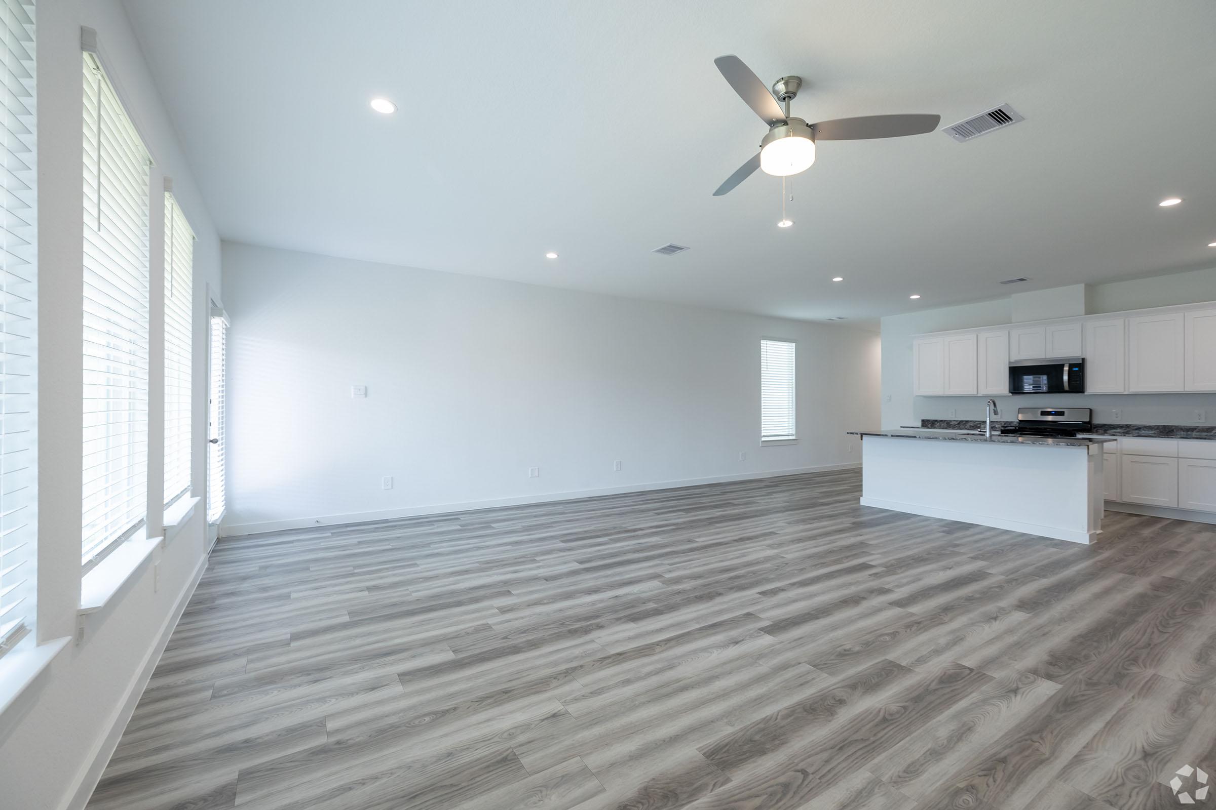 A spacious, modern living area featuring light-colored walls and wood-like flooring. Natural light streams in through large windows on one side. A ceiling fan is visible, and to the right, an open kitchen area with white cabinets and a countertop. The overall atmosphere is bright and airy.