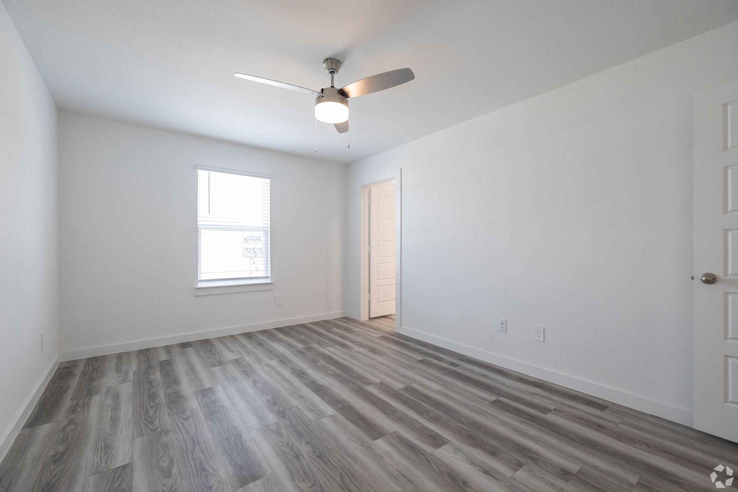 A bright, empty room with light gray walls and a ceiling fan. There's a window with white blinds letting in natural light, and wood-like flooring throughout. A white door leads to another area, creating a spacious and modern atmosphere.