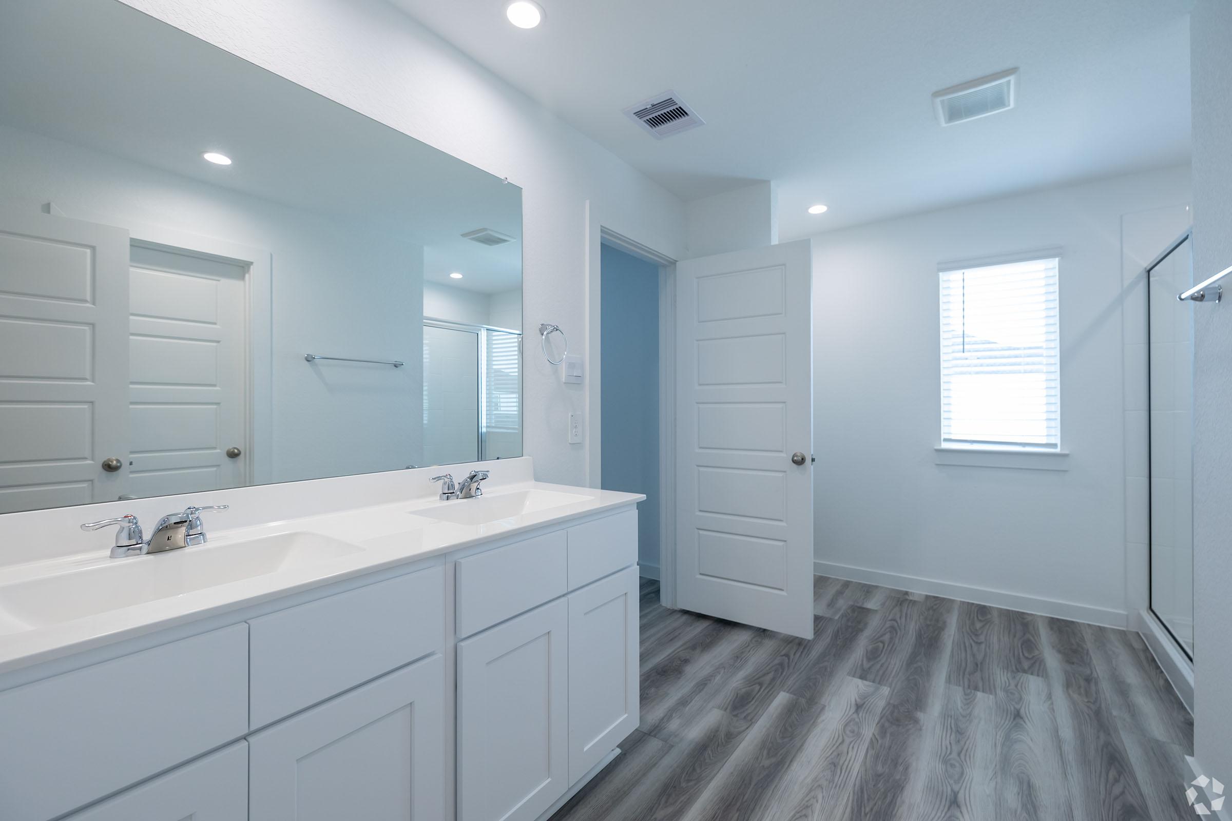 A modern bathroom featuring a double vanity with two faucets, a large mirror above, and neutral-colored walls. Natural light enters through a window. The floor is styled with wood-like laminate, and a glass shower is visible in the corner. A white door leads to another room.