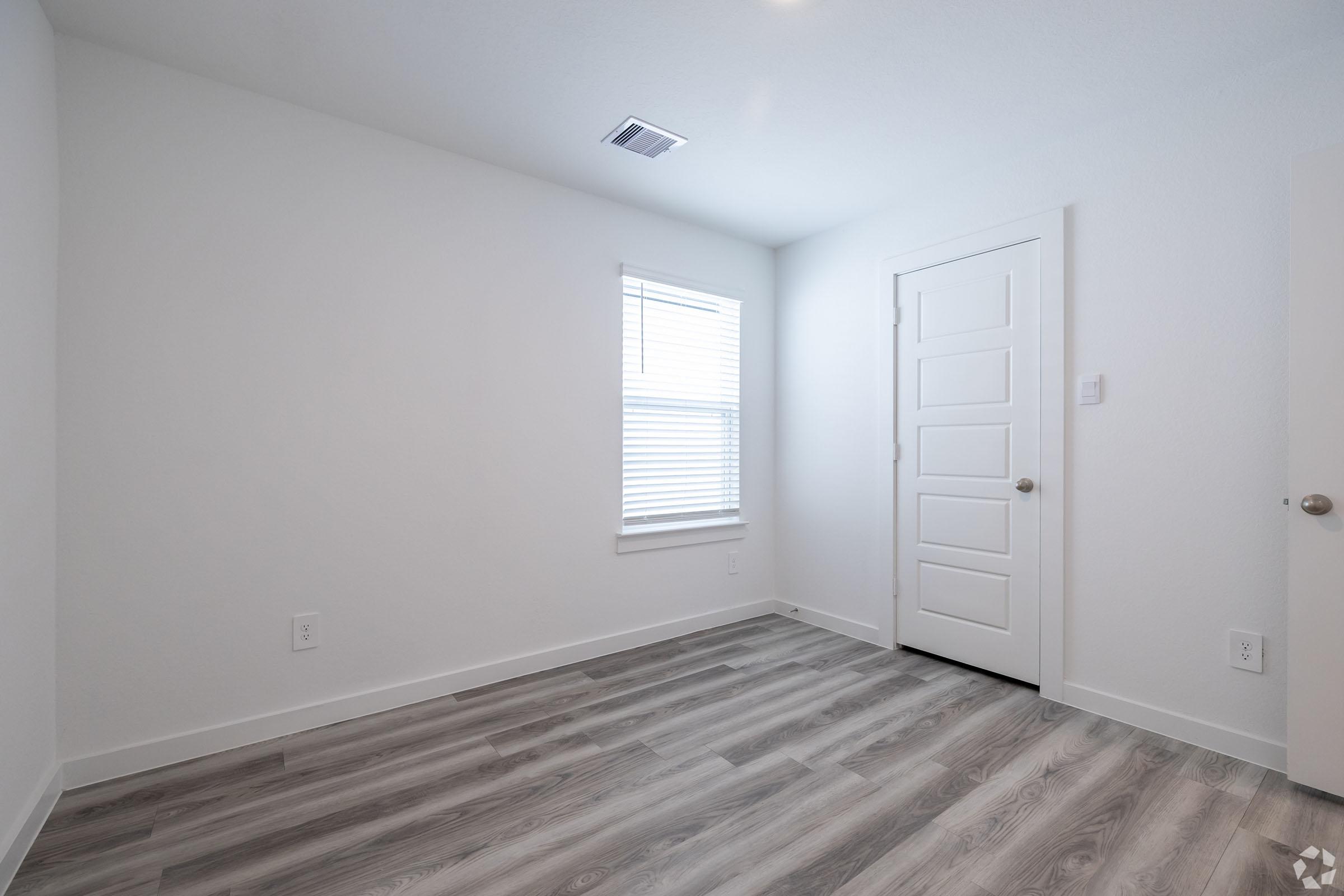 A bright, empty room with light-colored walls and grey laminate flooring. The room features a window with white blinds, allowing natural light, and a closed white door. The minimalist design provides a spacious feel, ideal for various uses.