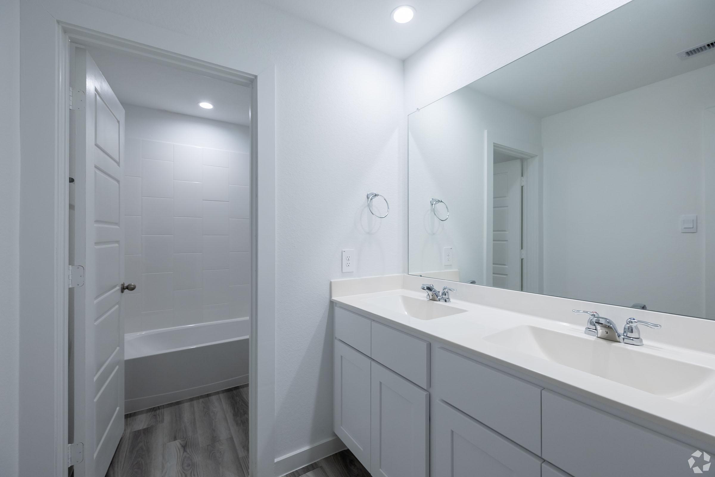 A modern bathroom featuring a double vanity with white sinks and mirrors, light gray walls, and vinyl flooring. A white bathtub is visible in a separate area behind a door, with stylish fixtures and natural lighting enhancing the clean, contemporary design.