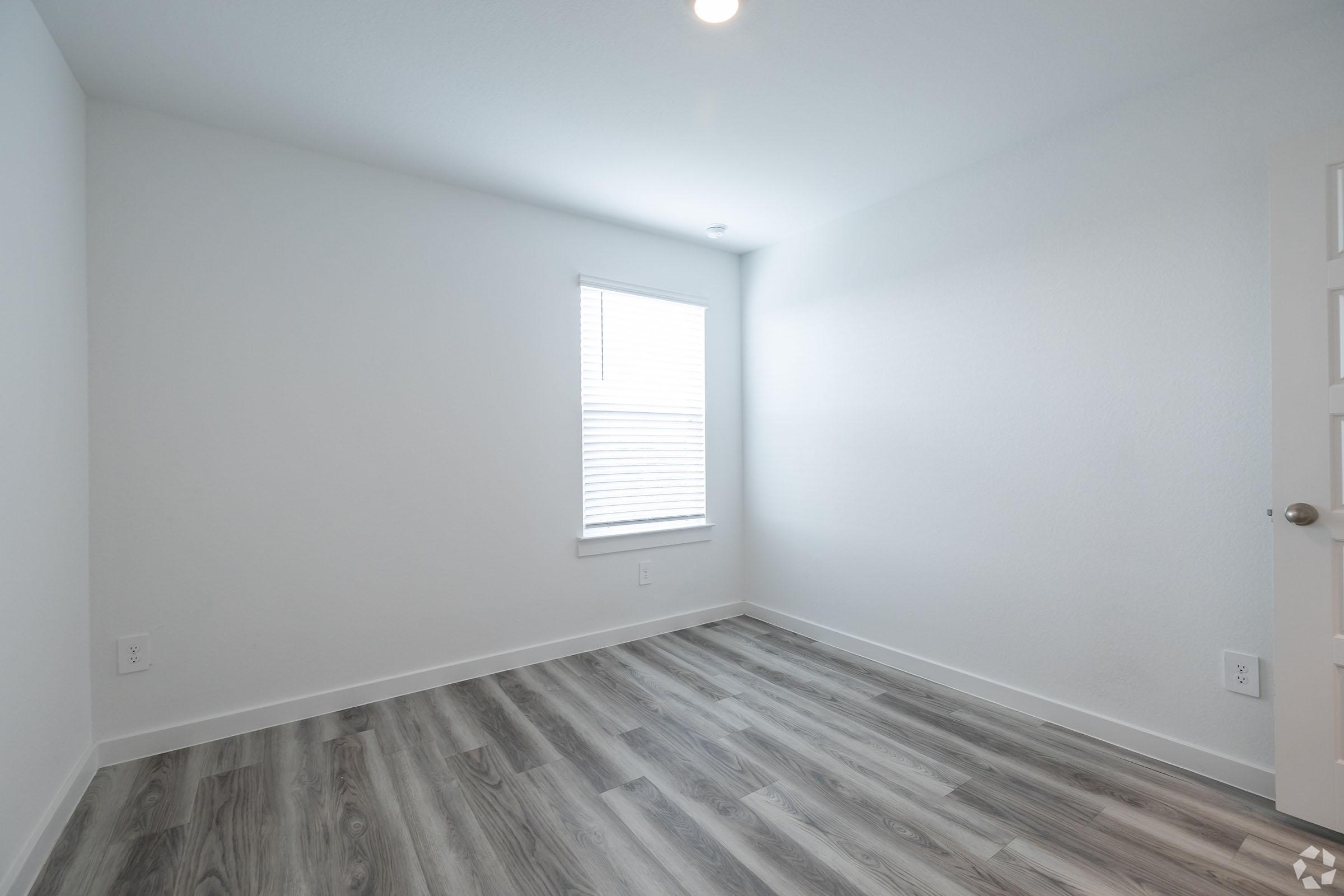 A bright, empty room with light gray walls and a window covered by white blinds. The floor features light wood-like laminate, and there is a white door on the right side. The overall ambiance is clean and minimalistic, ideal for personal decoration.