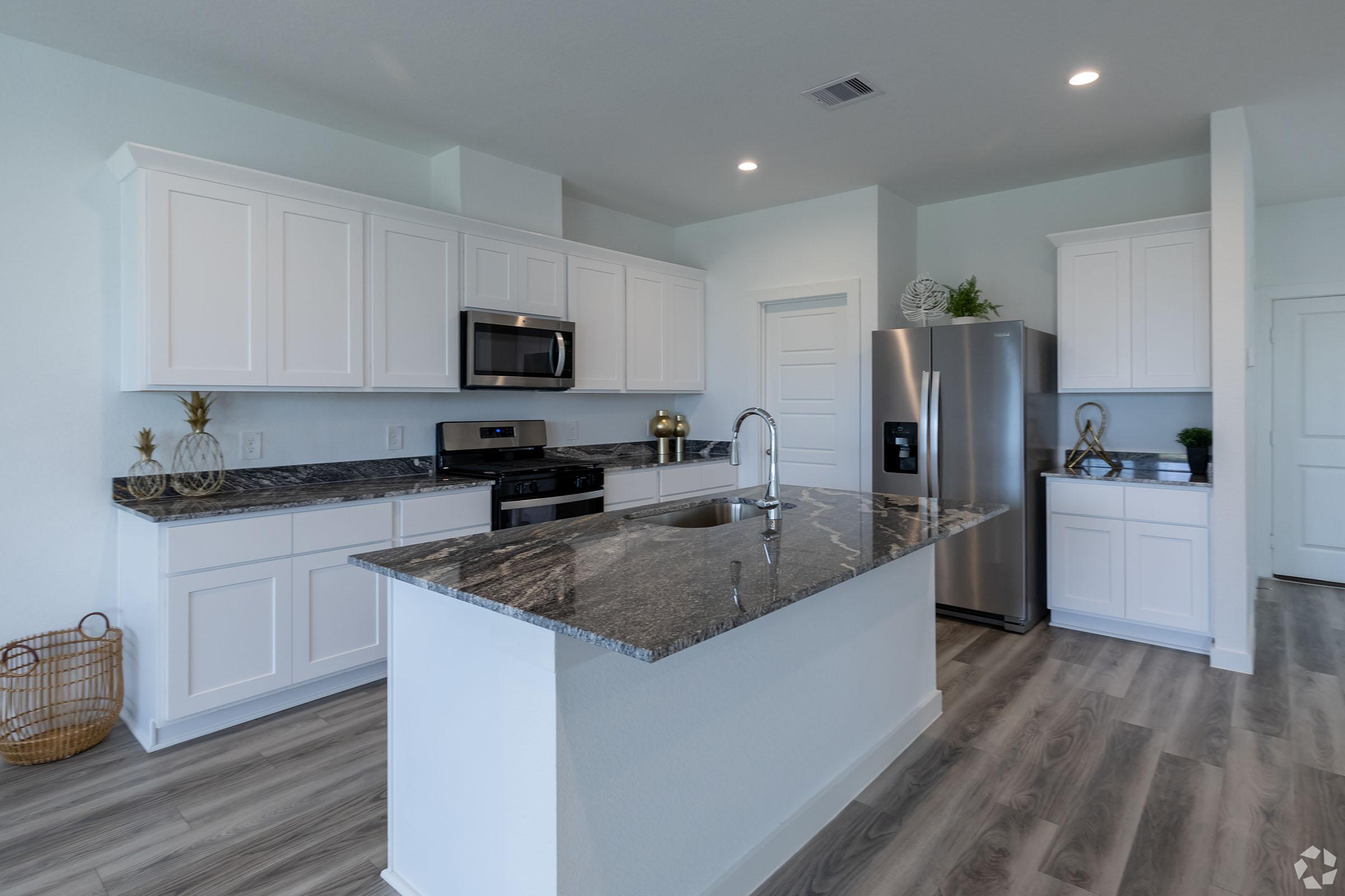 A modern kitchen featuring white cabinets, a large island with a dark granite countertop, stainless steel appliances including a microwave and refrigerator, and stylish decor. The open layout is bright with natural light, showcasing a clean and contemporary design.