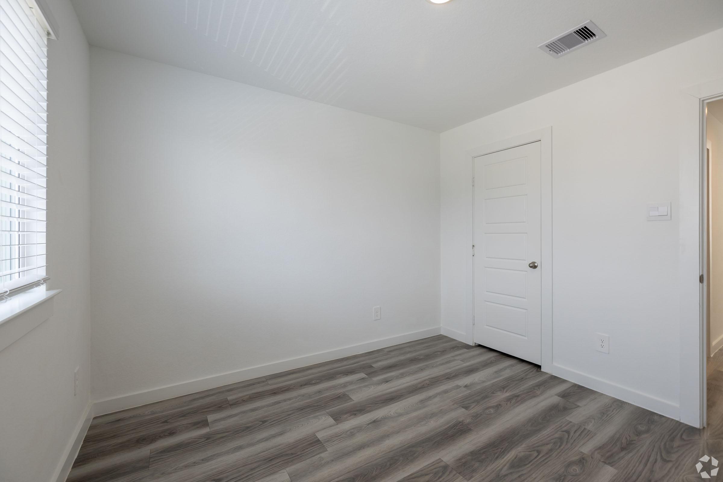 Empty interior room with light gray walls, a window with white blinds allowing natural light, and a closed white door. The floor is a light wood laminate. The room appears fresh and newly renovated, with no furniture present.