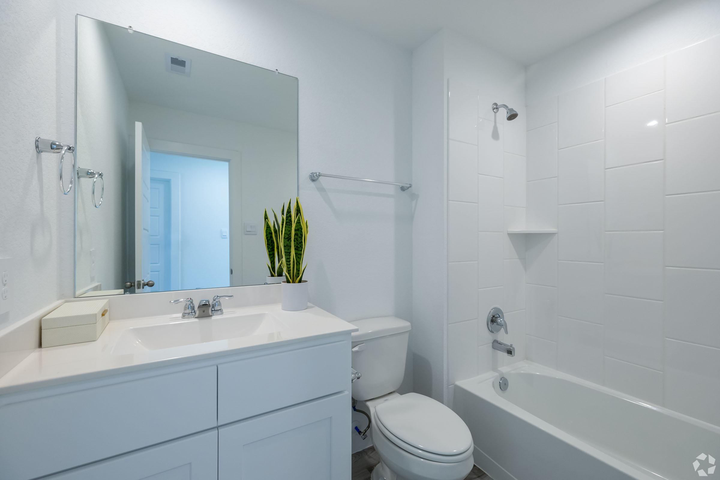 A modern bathroom featuring a white vanity with a mirror, a sink, and storage. The space includes a toilet, a bathtub with a showerhead, and light-colored walls. A potted plant adds a touch of greenery, and there is a door leading to another room in the background.