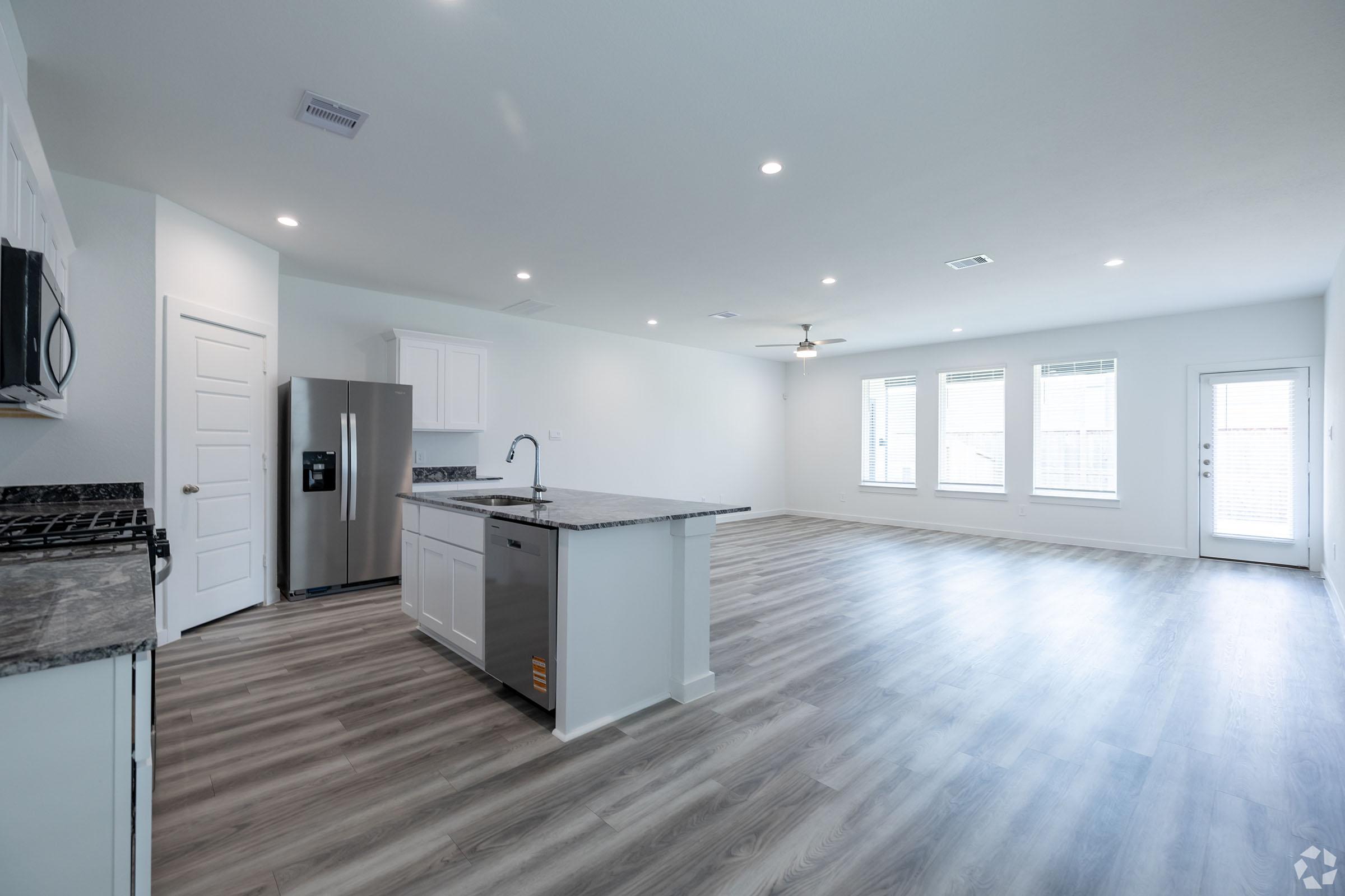 A modern, open-concept living space featuring a kitchen with a granite countertop island, stainless steel appliances, and white cabinetry. The area has ample natural light from large windows, a ceiling fan, and a patio door leading outside. The flooring is light-colored wood laminate.