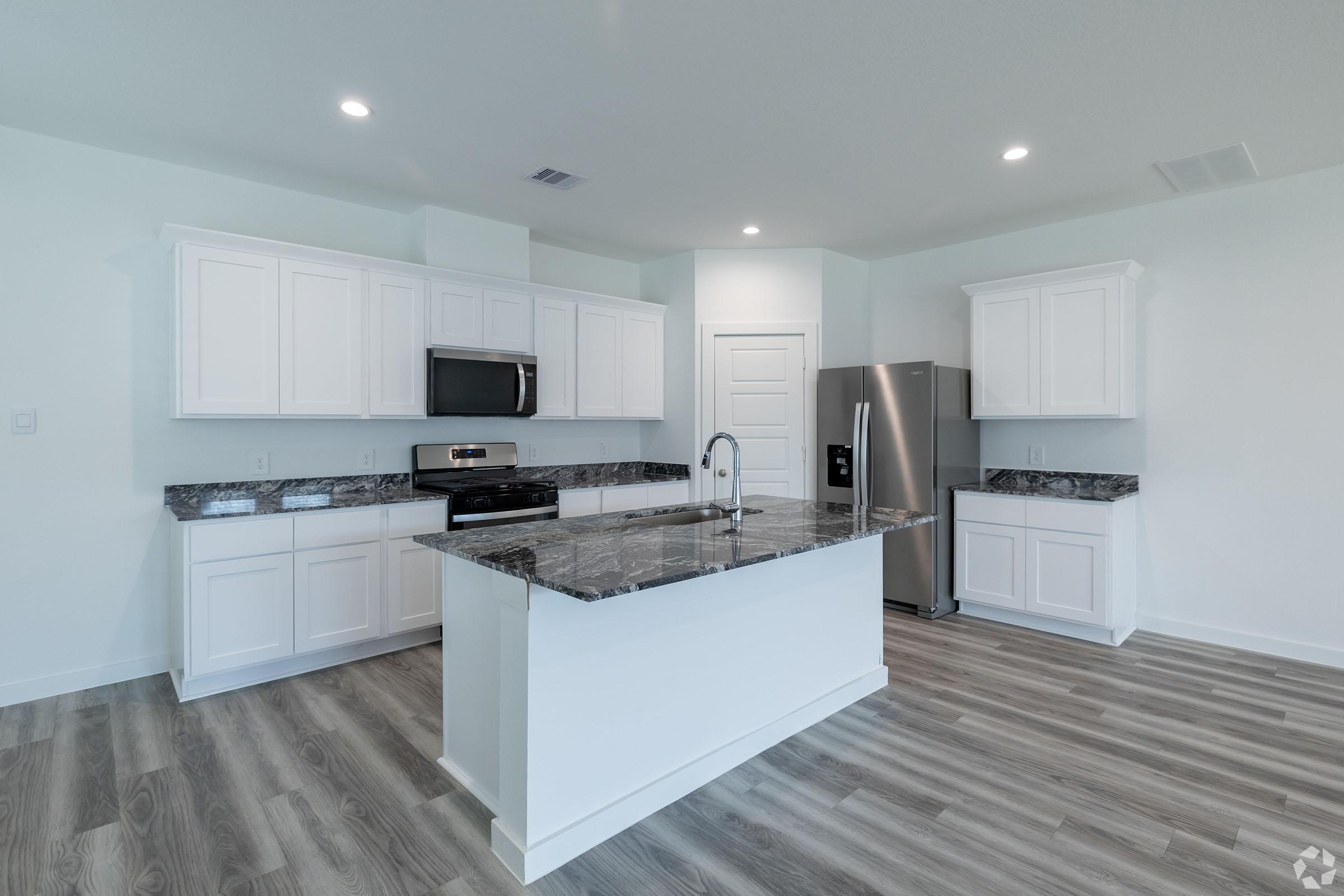 A modern kitchen featuring white cabinets, a granite countertop island with a sink, stainless steel appliances including a refrigerator and oven, and light-colored wood-like flooring. The space is bright and airy, designed with a clean, contemporary aesthetic.