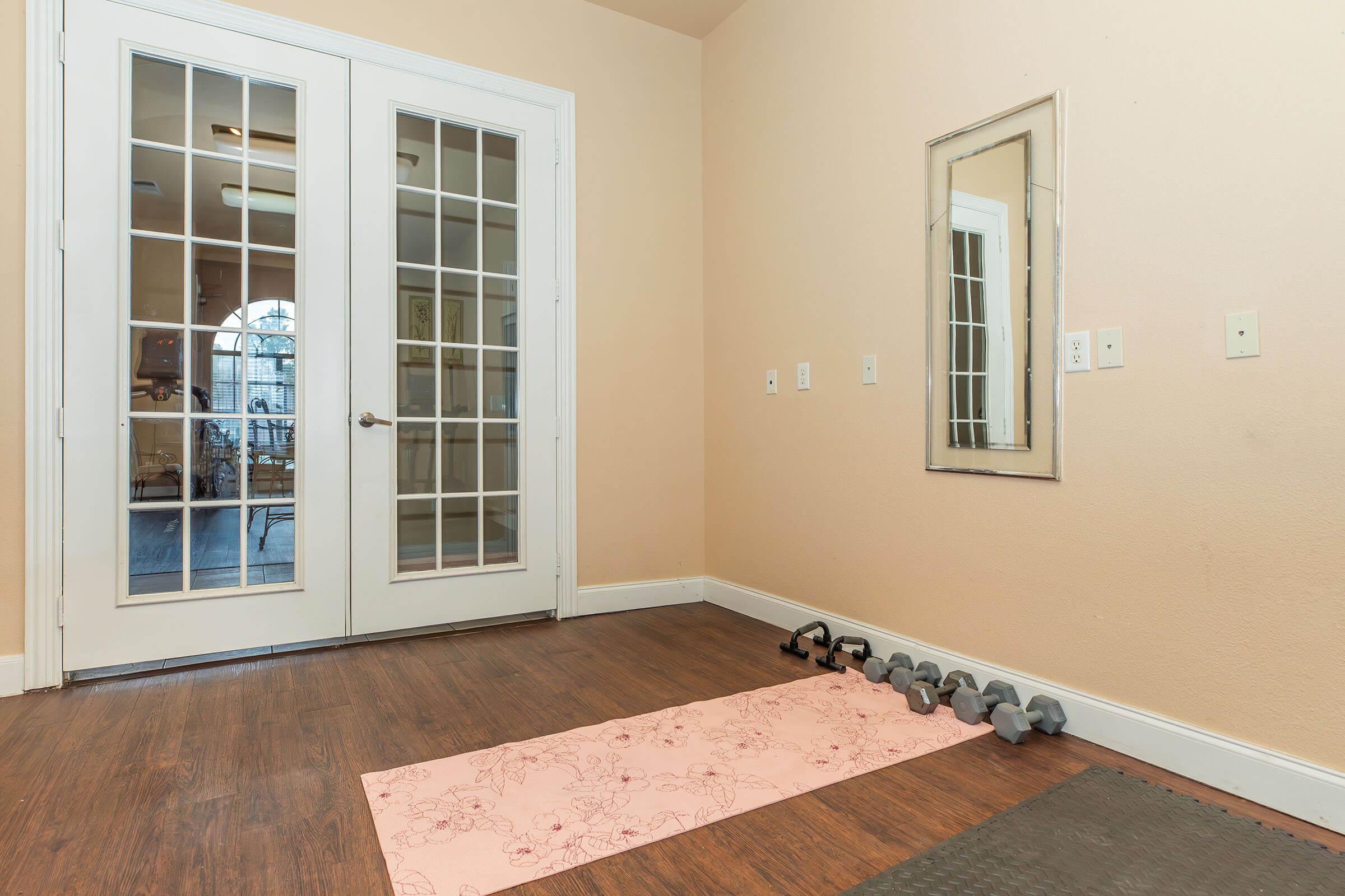 Home workout space featuring a light-colored wall, wooden flooring, a pink exercise mat on the floor, a set of dumbbells arranged nearby, a full-length mirror on the wall, and double glass doors leading to another room. The area is well-lit and organized, suitable for exercise and fitness activities.