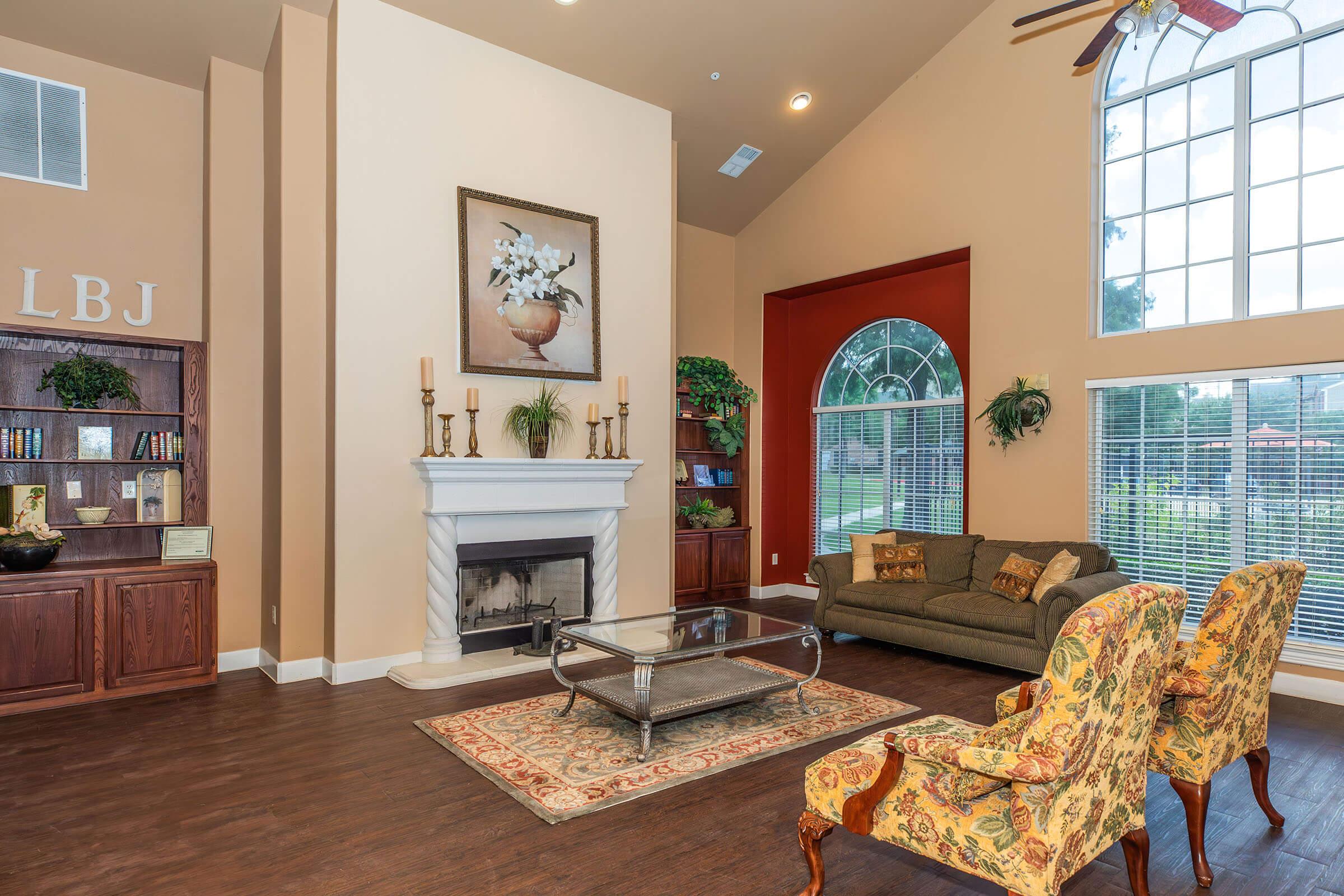 Cozy living room featuring a fireplace with a decorative mantle, a large window allowing natural light, a comfortable green sofa, and floral armchairs. A bookshelf with decorative items and plants adds warmth to the space, while a painting of flowers adorns the wall. The color palette is neutral and inviting.