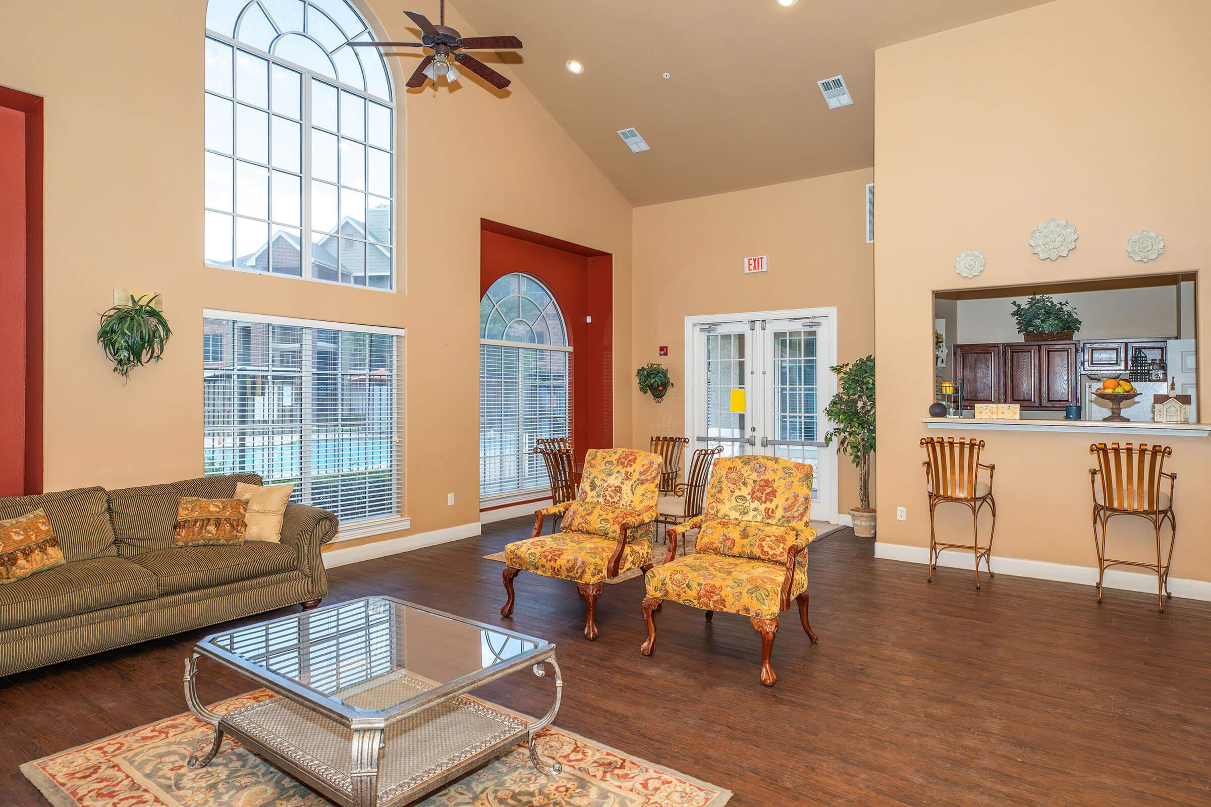 A spacious, well-lit living area featuring large windows, warm beige walls, and a high ceiling. The room includes a comfortable brown sofa, two ornate armchairs, a coffee table, and a rug. A reception area with bar stools is visible in the background, along with decorative plants and a welcoming atmosphere.