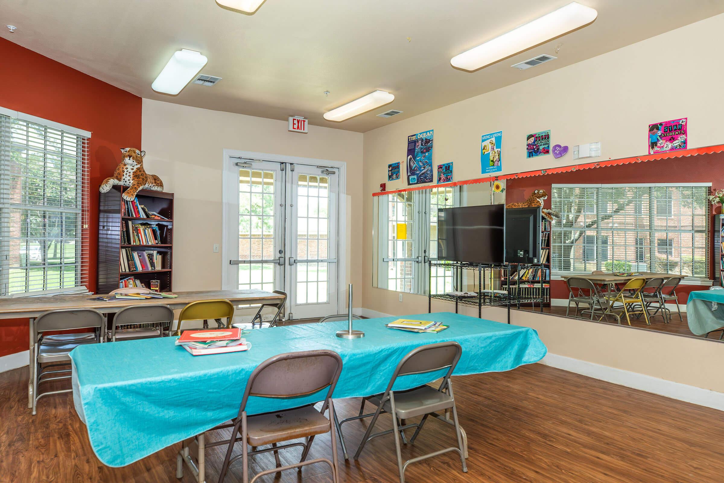 A bright and inviting room featuring a table covered with a turquoise tablecloth and several chairs arranged around it. There are bookshelves on one side, a mirror on the opposite wall, and colorful artwork displayed. Large windows allow natural light, and a television is visible on the wall.
