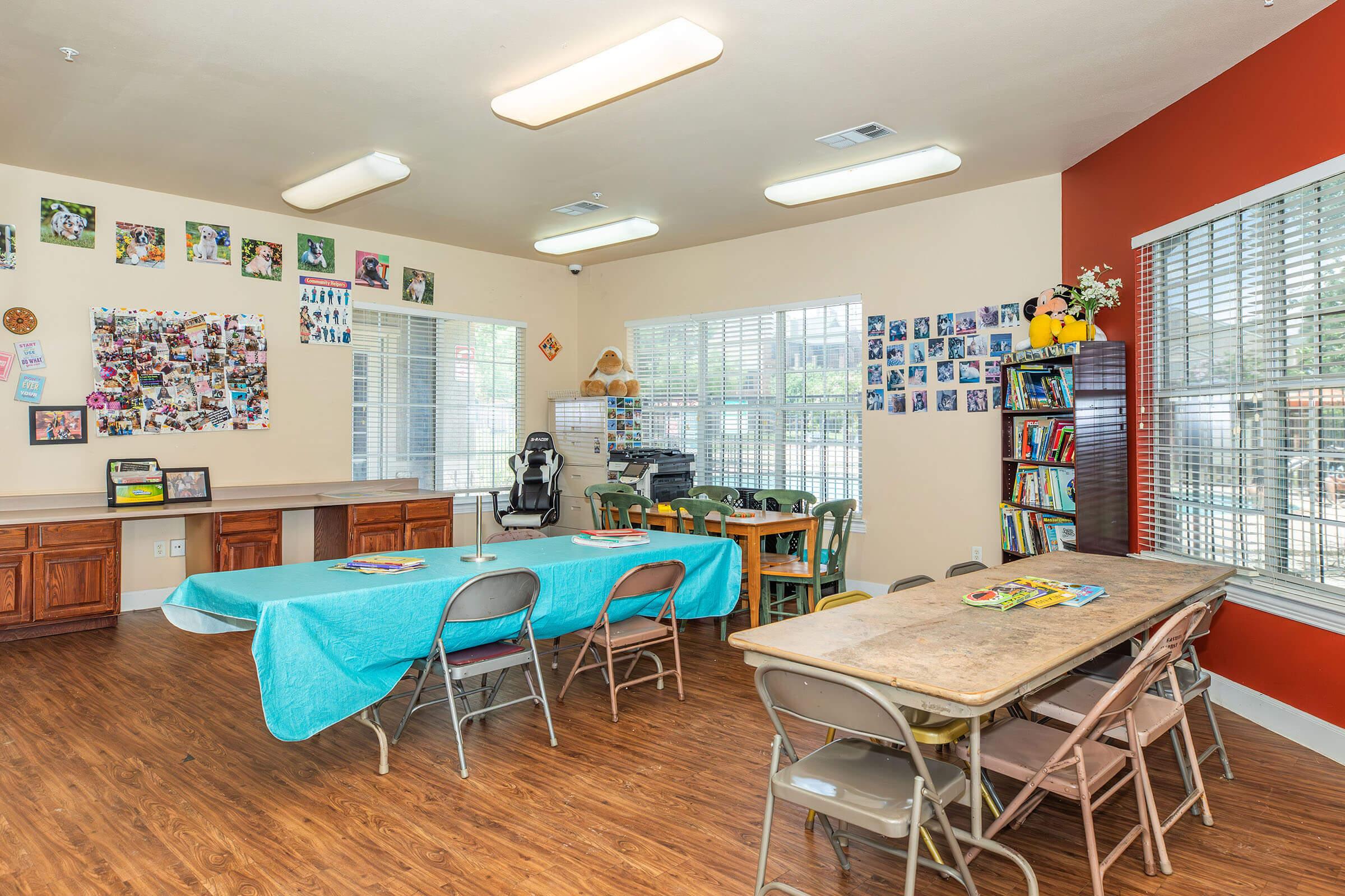 A spacious classroom featuring a blue table, multiple chairs, and wooden desks. Walls are adorned with various photos and artwork. Natural light streams through large windows, and a bookshelf is visible, filled with books. The room is well-organized and inviting, suitable for learning or group activities.