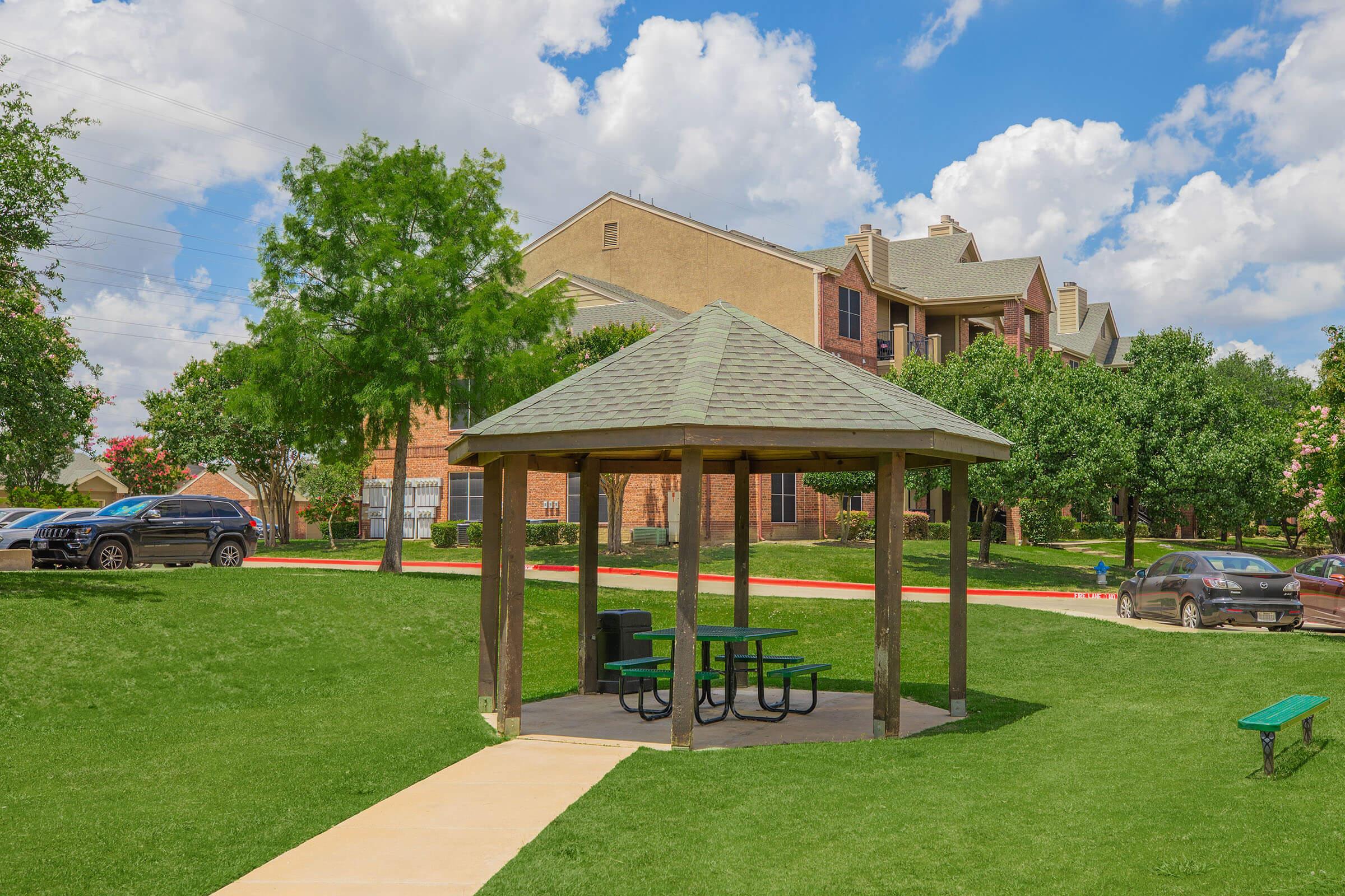 A gazebo with a sloped roof and green picnic tables is situated on a grassy area. Surrounding the gazebo are trees and landscaped grounds, with an apartment building in the background. The sky is partly cloudy, creating a bright and inviting atmosphere.