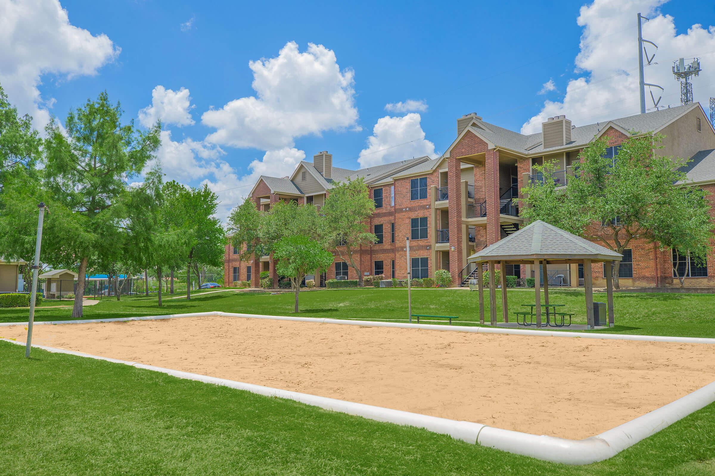 A sunny outdoor area featuring a sand volleyball court surrounded by green grass and trees. In the background, there's a large brick apartment building with multiple windows and a covered picnic area. Fluffy white clouds are visible in a bright blue sky.