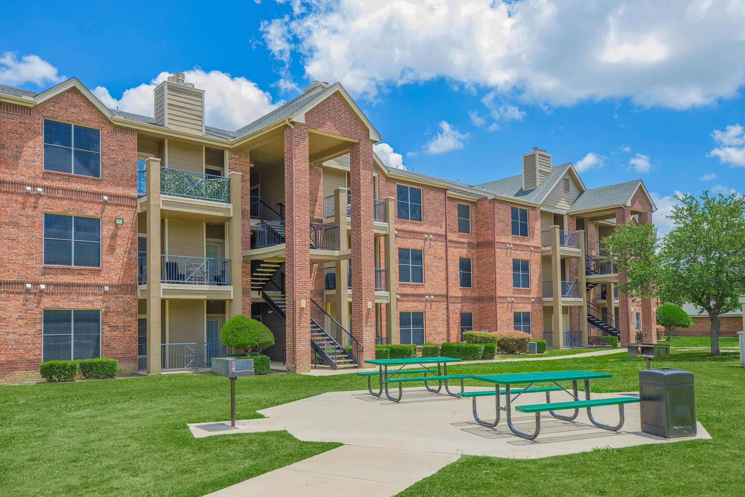 A modern brick apartment building with multiple levels, featuring balconies and staircases. In the foreground, there are green picnic tables on a concrete path, surrounded by well-maintained grass and small trees. The sky is blue with scattered clouds.