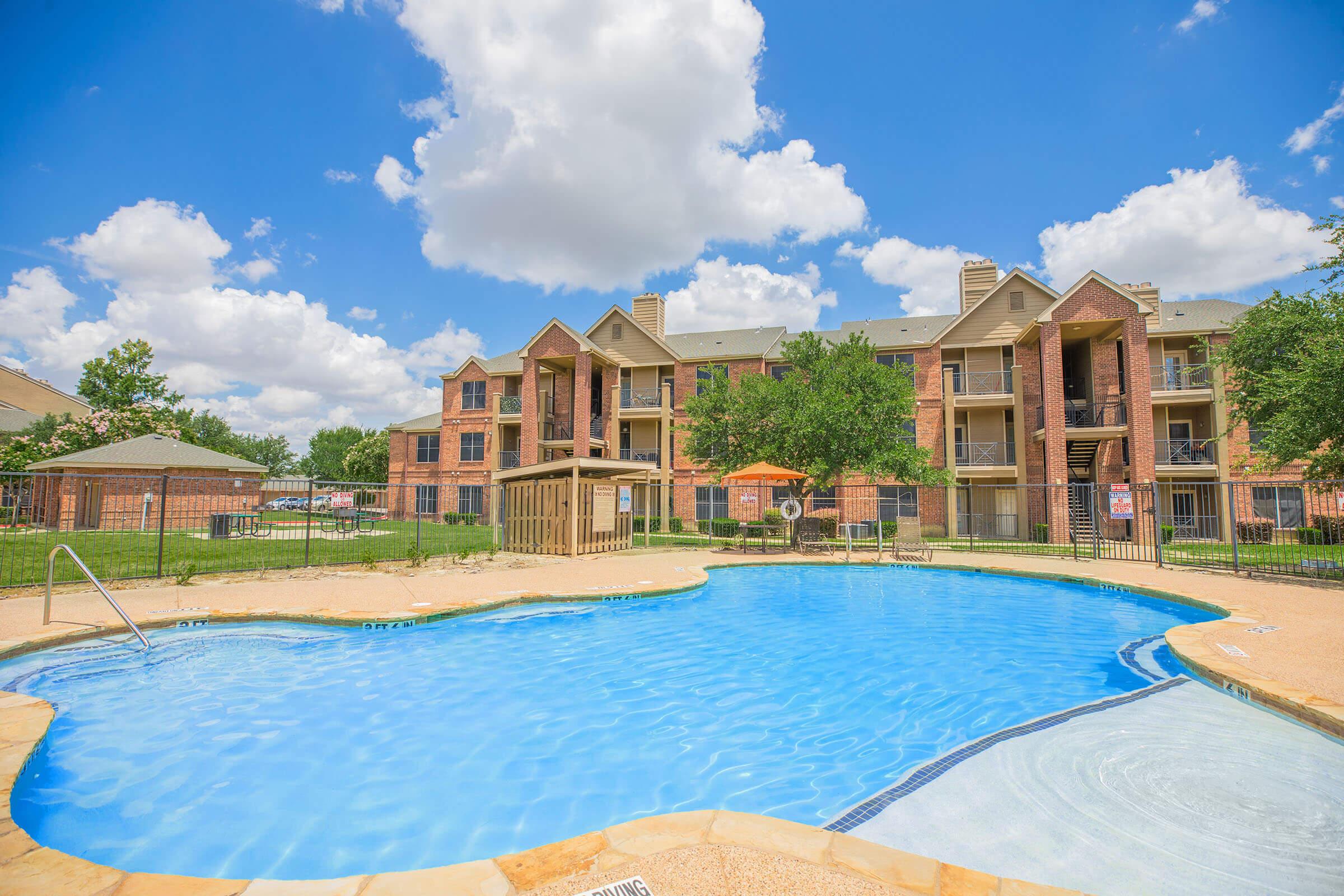 A sunny outdoor pool area with a large, blue swimming pool surrounded by a concrete deck. There are lounge chairs and an umbrella, with green grass and trees nearby. In the background, several brick apartment buildings are visible under a bright blue sky with fluffy white clouds.