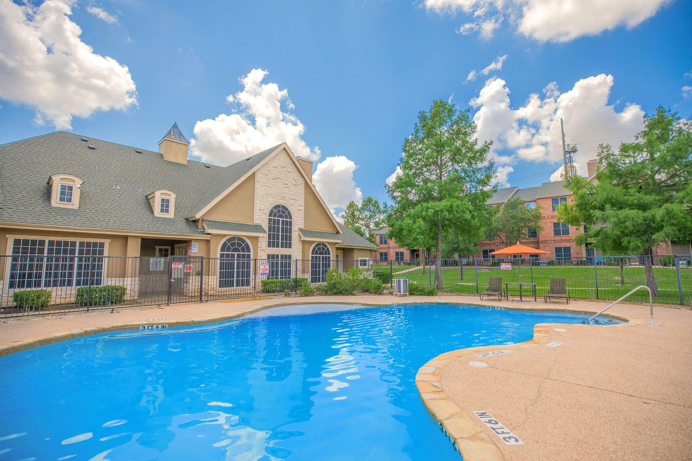 A clear blue swimming pool surrounded by a patio area, with lounge chairs and lush greenery. In the background, a large building with a stone facade and large windows is visible, along with additional buildings and a clear blue sky dotted with fluffy white clouds.