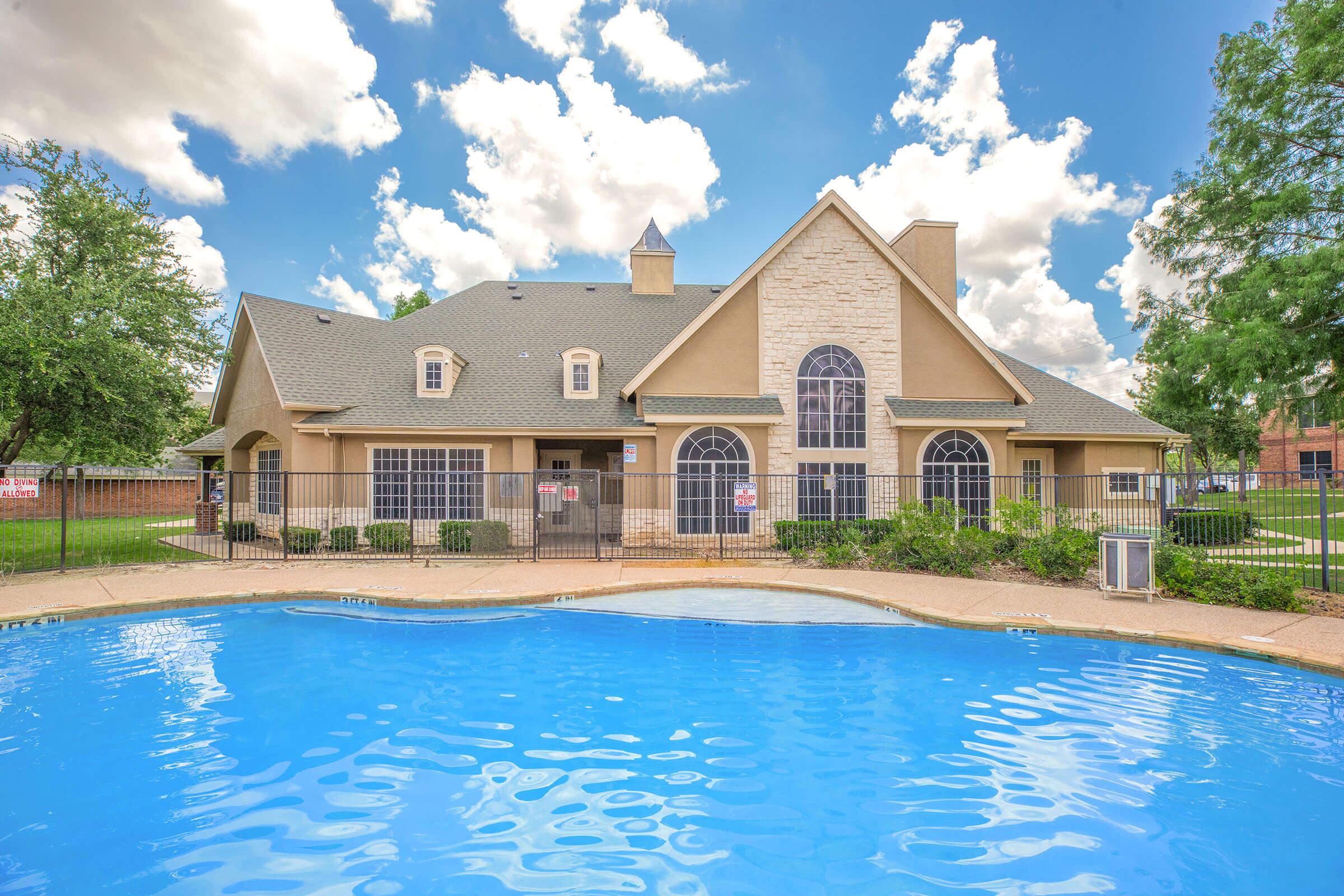 A modern building with a peaked roof and large windows next to a swimming pool. The exterior features light-colored stone and tan siding. Lush green landscaping surrounds the pool area, and the sky is bright with fluffy white clouds. The atmosphere is inviting and cheerful, suggesting a relaxing environment.